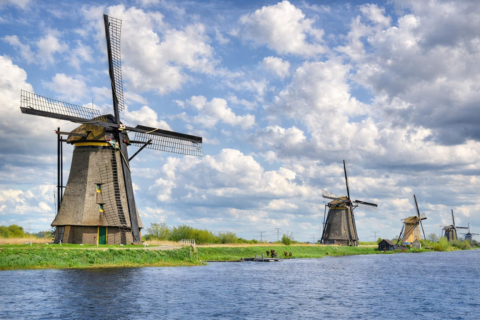 Weite Landschaft von Kinderdijk mit historischen Windmühlen entlang eines ruhigen Kanals, grünen Deichen und einem kleinen Steg im Vordergrund, unter hellem Sonnenlicht und einem Himmel voller plastischer, leicht veränderter Wolkenformationen
