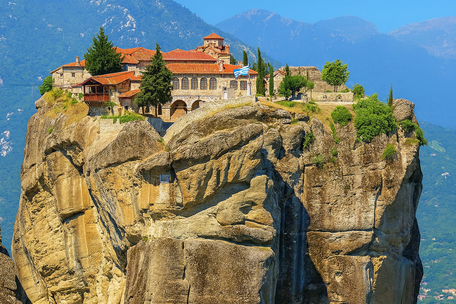 Das Kloster Agía Triáda in Meteora, Griechenland, thront auf einem hohen Felsen bei klarem Wetter und bestem Sonnenlicht vor den grünen Bergen von Thessalien.