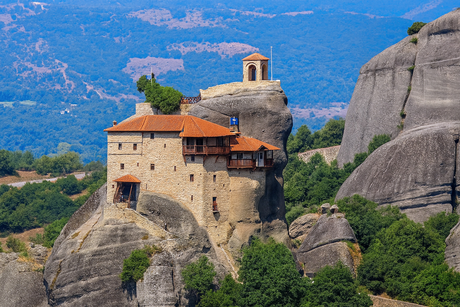 Das Kloster Agios Nikólaos in Meteora, Griechenland, thront auf einem steilen Felsen bei klarem Wetter und bestem Sonnenlicht, umgeben von grünen Hängen und Felsformationen.