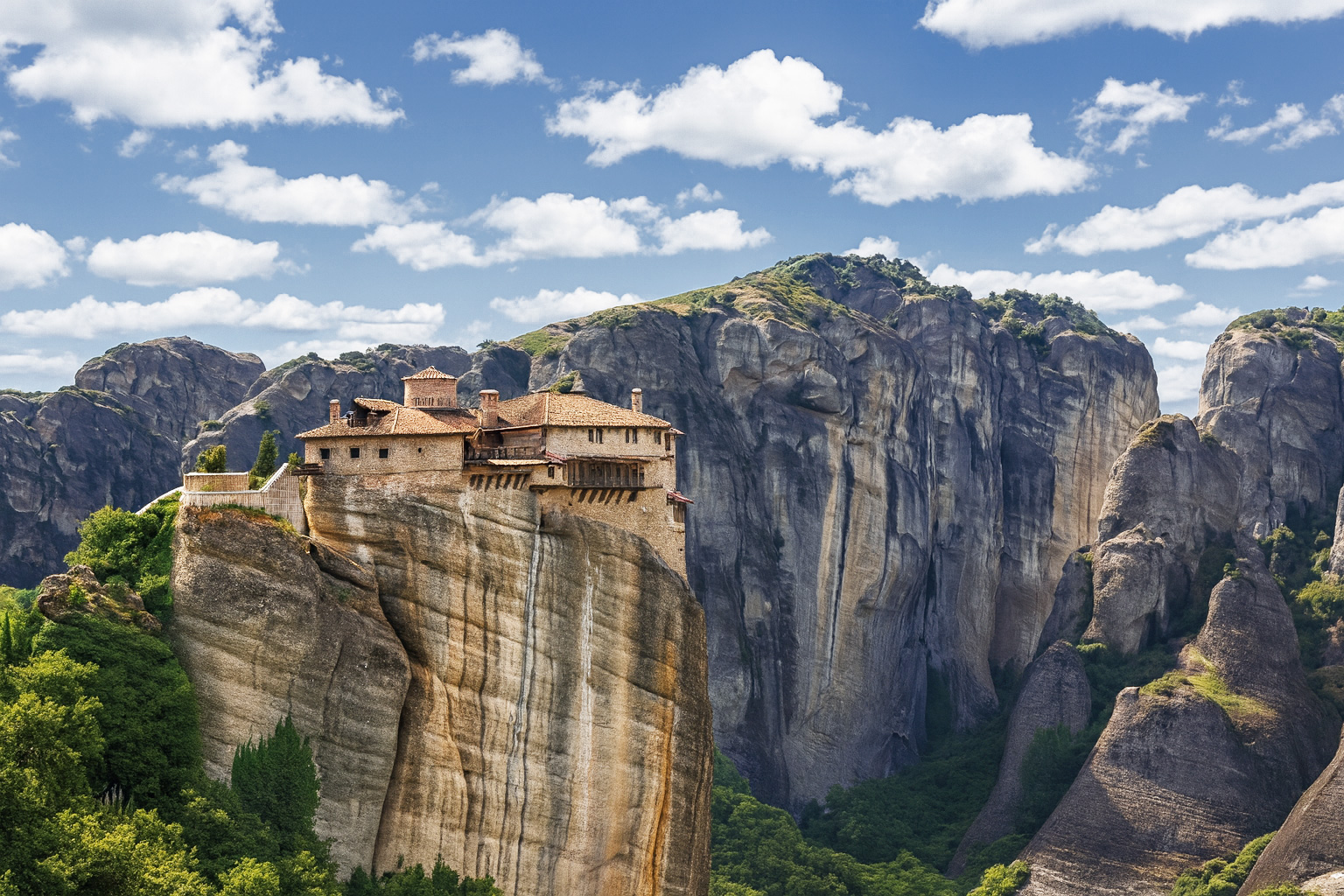 Das Kloster Rousánou in der Meteora-Region in Griechenland thront auf einem hohen Felsen bei Sonnenschein, umgeben von imposanten Felsformationen und malerischen weiß-grauen Wolken