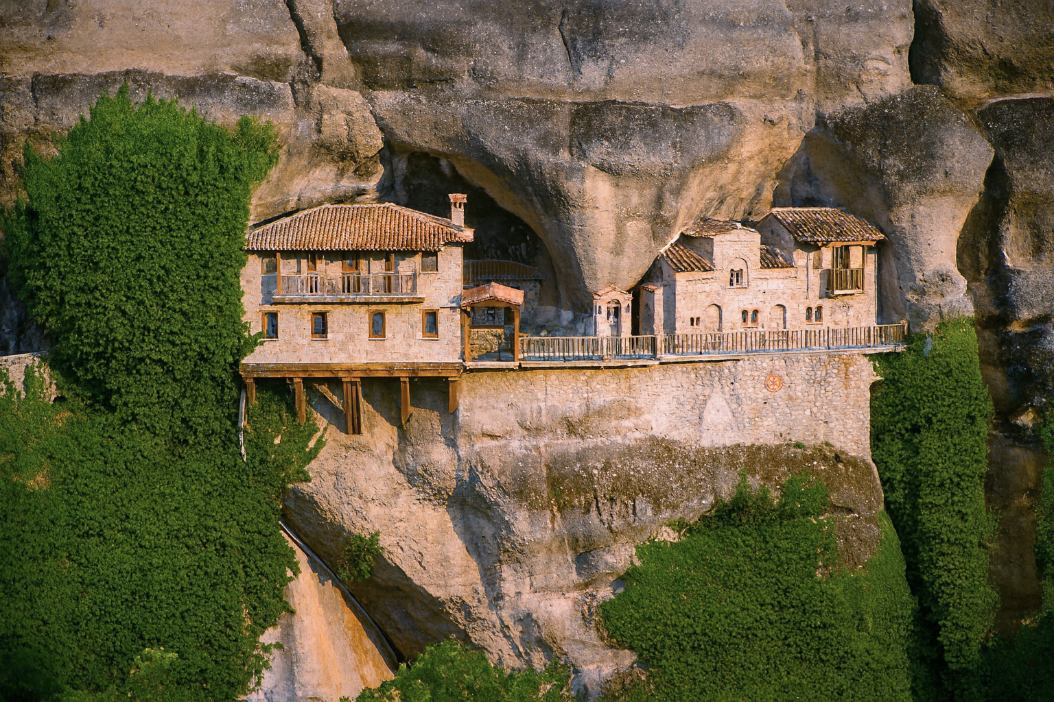 Das Kloster Ypapantí in Meteora, Griechenland, liegt in einer Felsnische bei klarem Wetter und bestem Sonnenlicht von rechts, umgeben von grün bewachsenen Felsen.