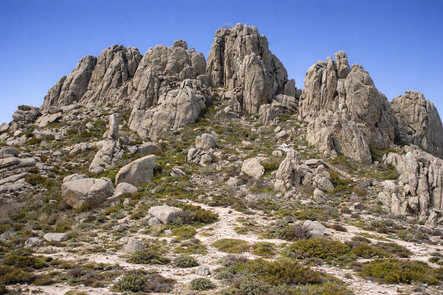 Granitlandschaft von La Pedriza mit bizarr geformten Felsformationen und zerklüfteten Türmen, dazwischen ein schmaler Wanderpfad durch niedrige Vegetation und Geröll im kräftigen Sonnenlicht unter klarem blauem Himmel