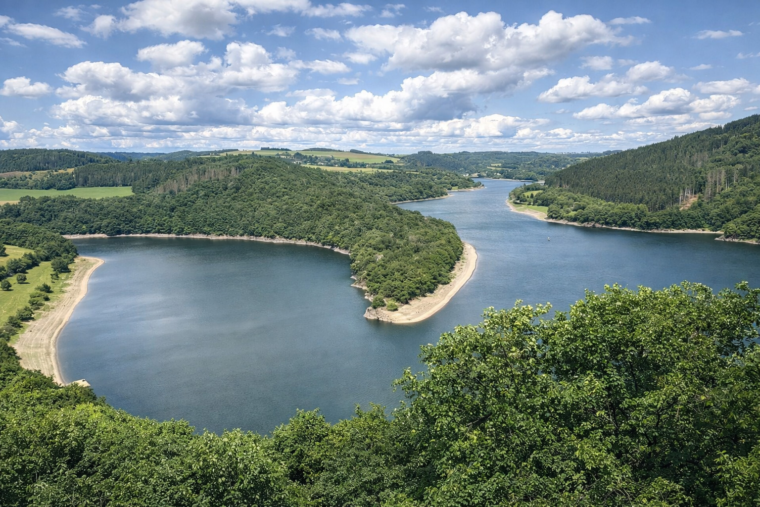 Panoramablick auf den Lac de la Haute-Sûre in Luxemburg, der sich in breiten Kurven durch bewaldete Hügel schlängelt, mit hellen Uferbänken und ruhiger Wasseroberfläche, im Vordergrund dichtes Blattgrün, darüber ein Himmel mit leicht veränderten Quellwolken im klaren Sonnenlicht