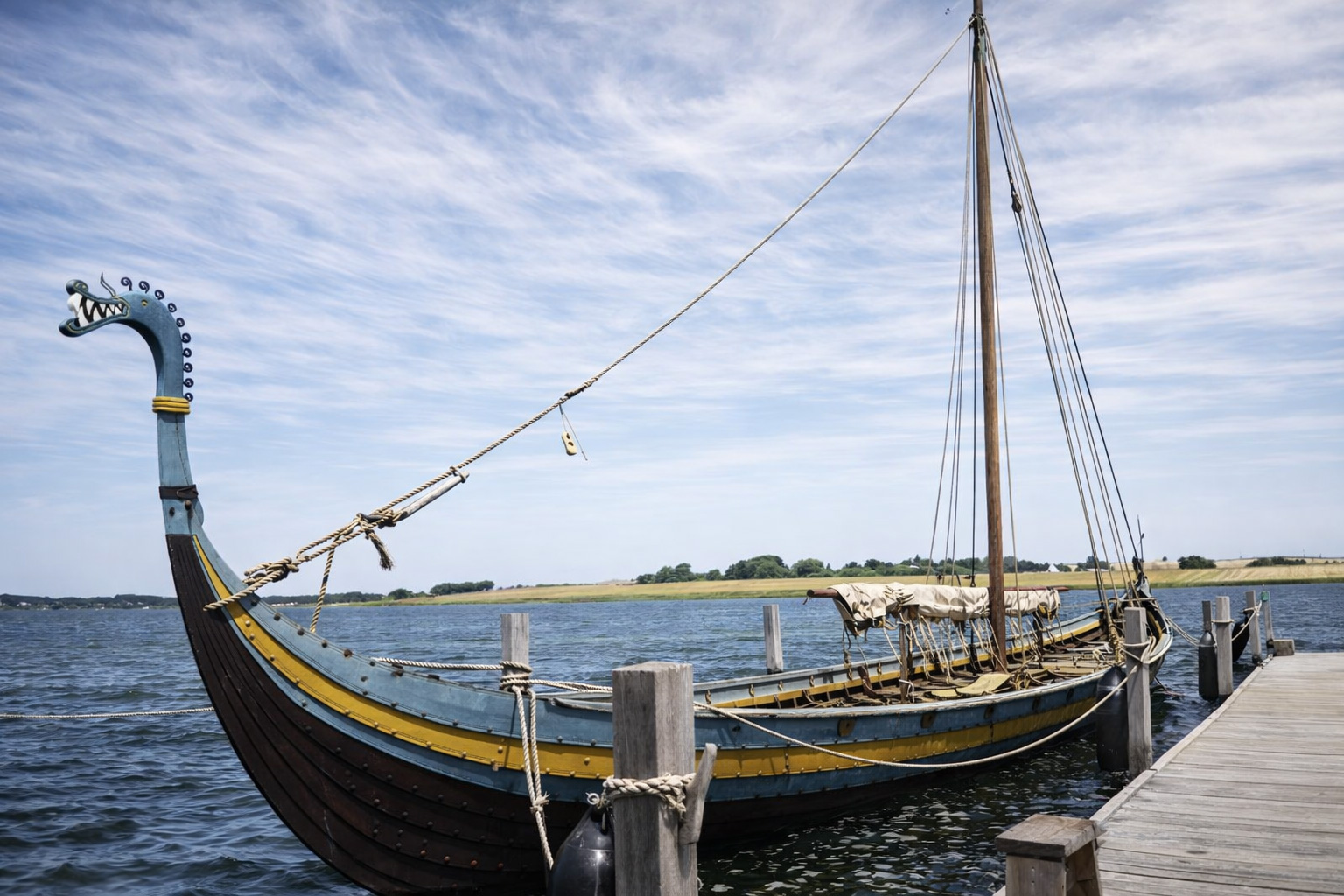 Wikingerschiff mit Drachenkopfbug am Steg des Ladby Vikingemuseum, das Holzboot liegt vertäut im ruhigen Wasser neben einem hölzernen Anleger, im Hintergrund flache Küstenlandschaft und Felder, darüber ein weiter Himmel mit feinen, nebligen Zirruswolken