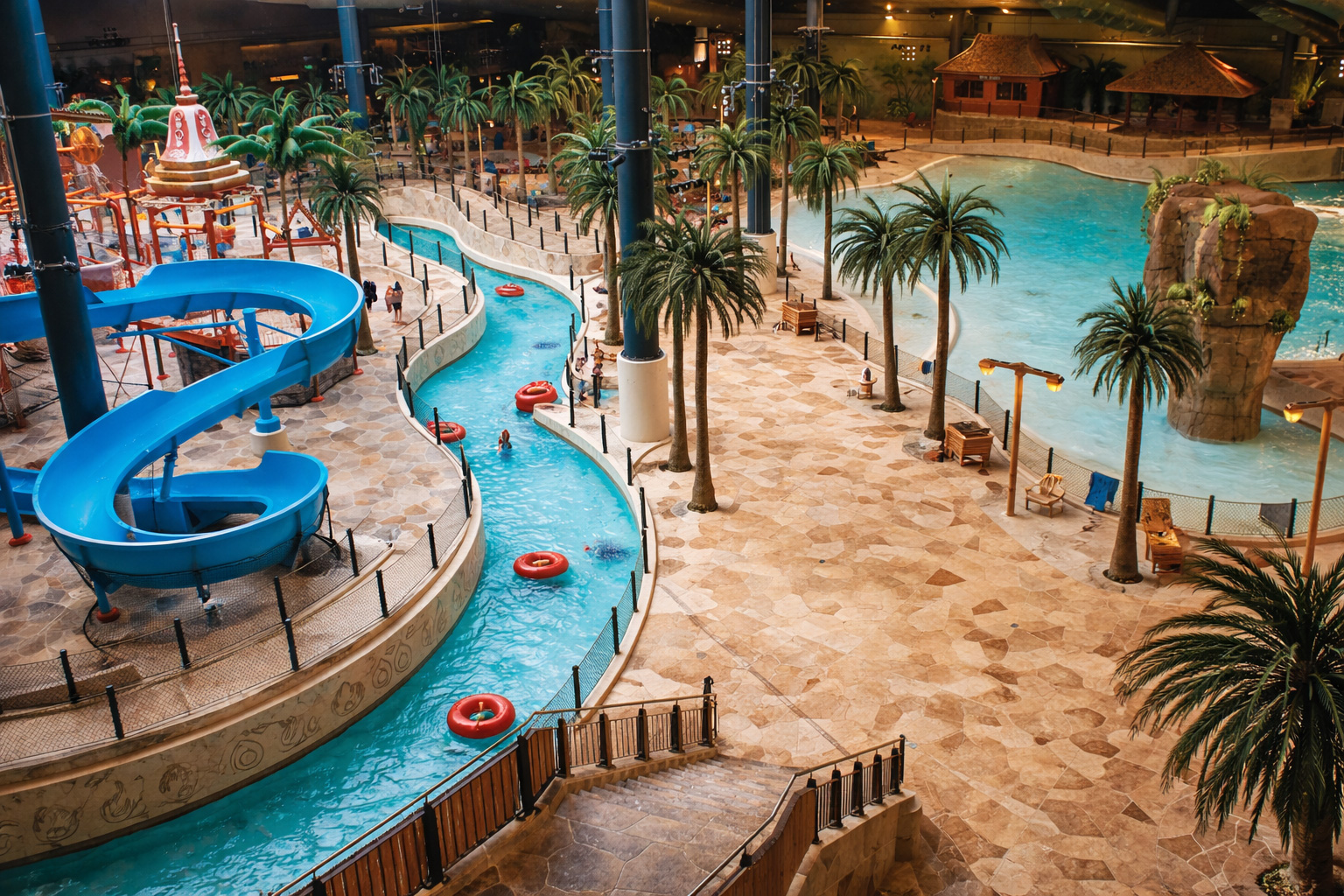 Blick von oben in das tropisch gestaltete Erlebnisbad Lalandia Billund mit türkisfarbenem Lazy River, roter Schwimmreifenstrecke und großer blauer Rutsche links, Palmen und Liegebereiche auf heller Steinfläche, rechts ein weitläufiges Wellenbecken mit Felsinsel und strohgedeckten Hütten im Hintergrund