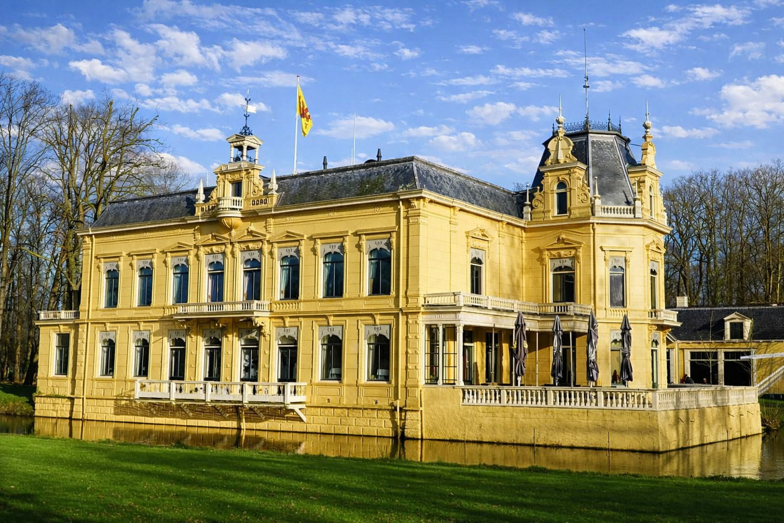 Historisches gelbes Herrenhaus Landgoed Nienoord mit reich verzierten Fassaden, Balkon und Türmchen, umgeben von gepflegtem Rasen und Wassergraben, aufgenommen bei klarem Sonnenlicht mit leicht veränderten Wolken am Himmel