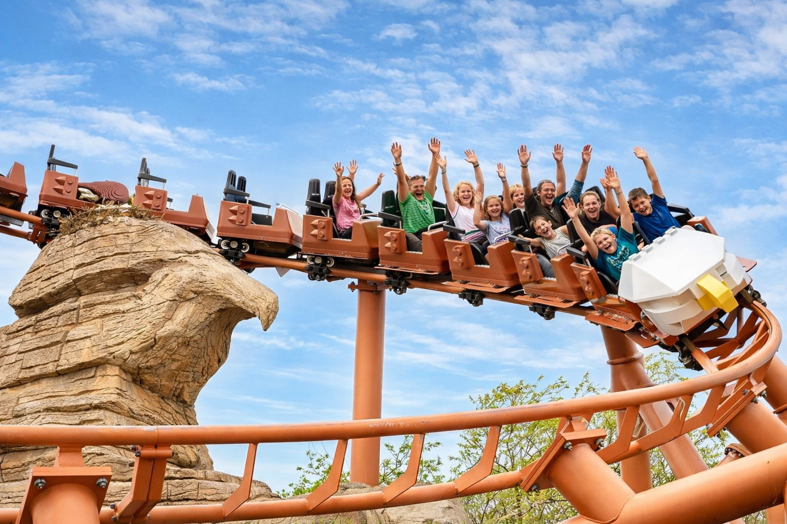 Achterbahn im LEGOLAND Billund mit orangefarbener Schienenkonstruktion und einem Wagen mit großer LEGO-Adlerfront, Besucher sitzen mit erhobenen Armen in den Sitzen, links eine Felskulisse in Form eines Adlerkopfs, darüber blauer Himmel mit leicht veränderten Wolken bei hellem Sonnenlicht und klaren Schatten