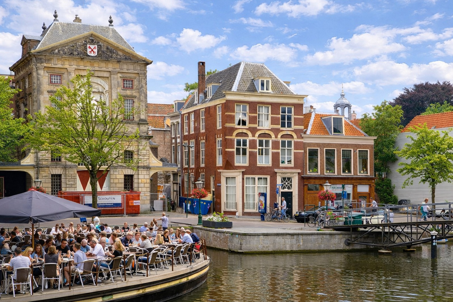 Kanalansicht in Leiden mit historischen Backstein- und Sandsteingebäuden am Ufer, kleiner Brücke und ruhigem Wasser mit Spiegelungen, links ein belebtes Café auf einer Plattform mit vielen Gästen unter Sonnenschirmen, darüber ein leicht bewölkter Himmel im klaren Sonnenlicht