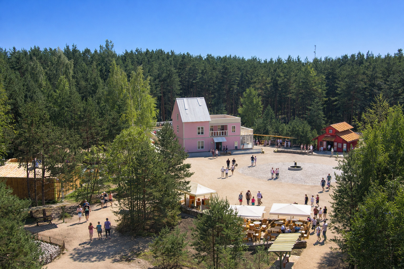 Luftaufnahme des Lottemaa Freizeitparks mit hellem Platz zwischen hohen Kiefern, einem rosa Gebäude und roten Holzhäusern, Sitzbereich mit Sonnenschirmen im Vordergrund, Besuchergruppen schlendern über den Platz, strahlender Sonnenschein mit klaren Schatten unter tiefblauem Himmel