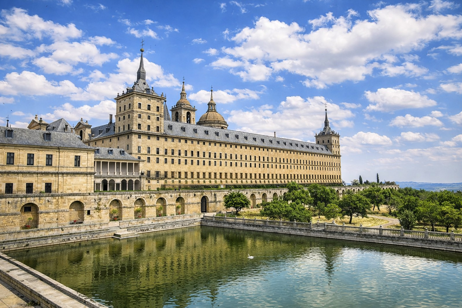 Weitblick auf das Monasterio de El Escorial mit langgestreckter Renaissancefassade, Türmen und Kuppeln, davor ein rechteckiges Wasserbecken mit Spiegelung und steinernen Arkaden, umgeben von Gärten und Hügeln unter leicht veränderten, lockeren Wolken