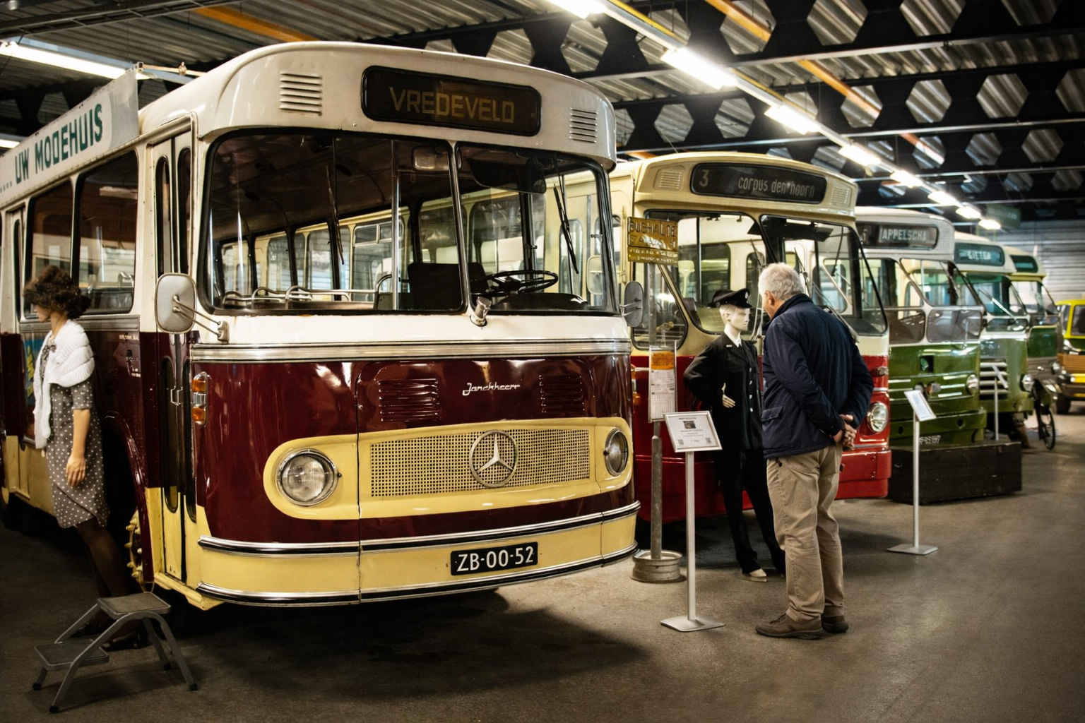 Innenansicht im National Bus Museum mit einer Reihe restaurierter historischer Busse unter Hallenbeleuchtung, im Vordergrund ein zweifarbiger Oldtimerbus mit großer Frontscheibe und Mercedes-Emblem, daneben Informationstafeln und ein Museumsgast, der die Ausstellung betrachtet