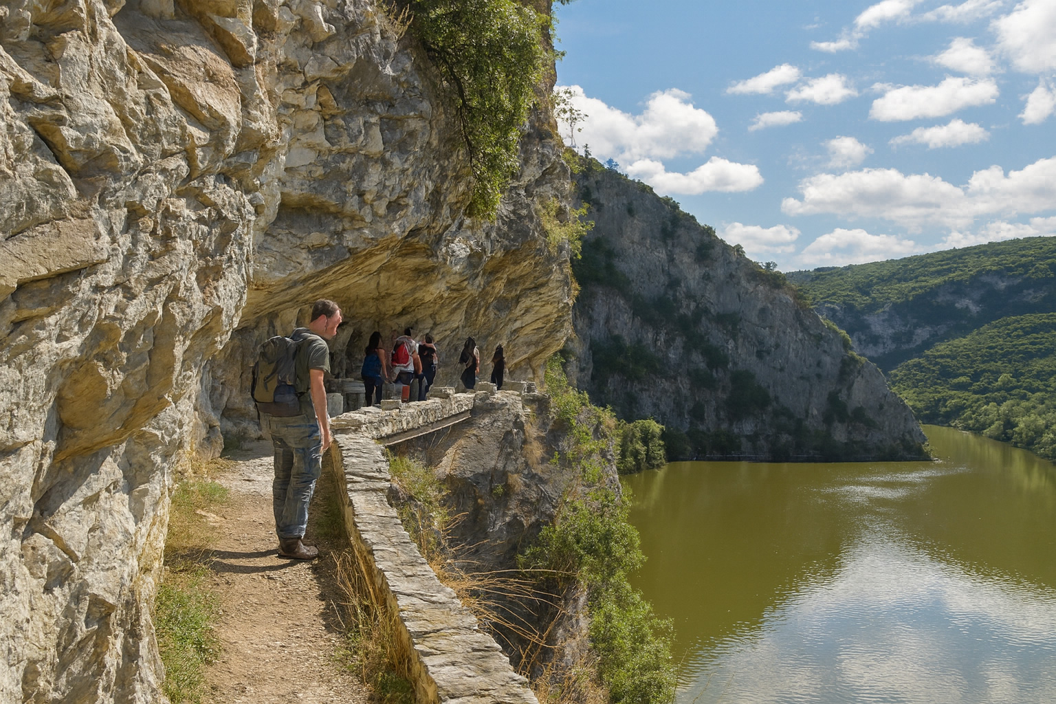Wanderer auf einem schmalen Felsenpfad über dem Fluss im Nestos Adventure Park in Griechenland, bei sonnigem Wetter mit weiß-grauen Wolken und grüner Landschaft im Hintergrund.
