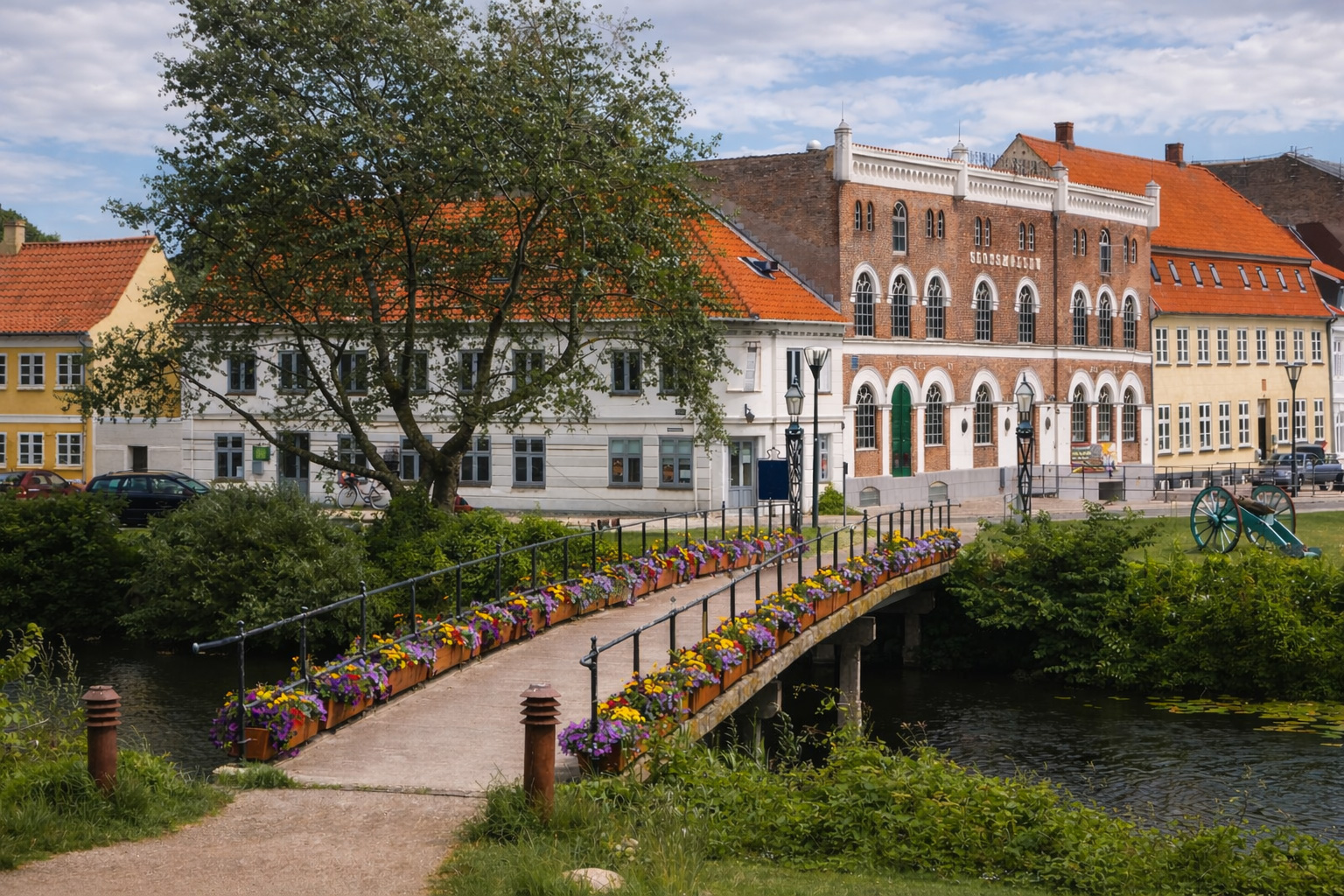 Blumengeschmückte Fußgängerbrücke über einen ruhigen Kanal in Nyborg, im Vordergrund bunte Blumen in Kästen entlang des Geländers, dahinter historische Gebäude mit roten Ziegeldächern und eine markante Backsteinfassade am Ufer, umgeben von Grünflächen und einem großen Baum.