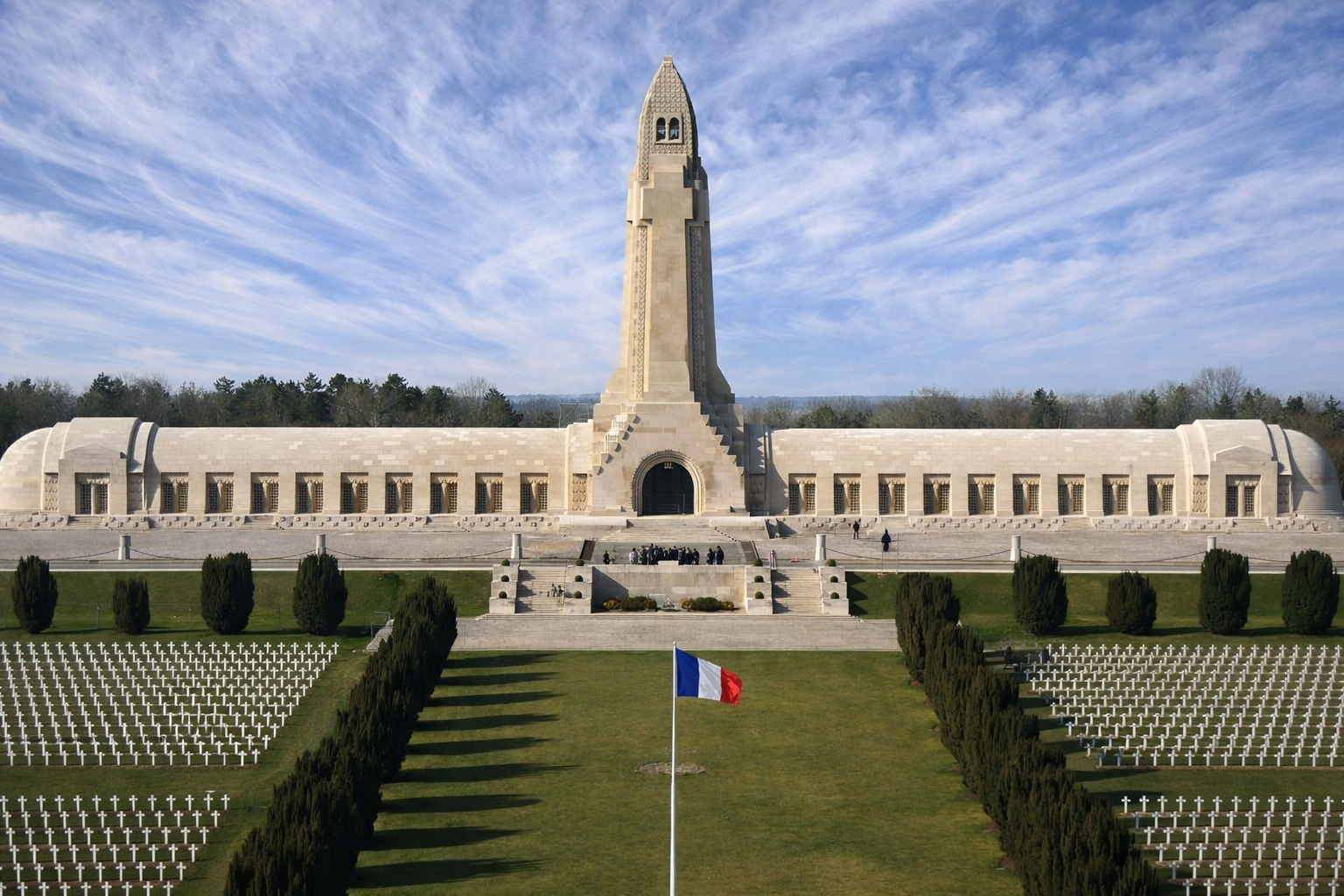 Monumentale Fassade des Ossuaire de Douaumont mit hohem Turm und langen Flügelbauten, davor Reihen weißer Grabkreuze und dunkle Zypressen entlang einer Rasenachse mit französischer Flagge unter einem hohen Himmel mit feinen Schleierwolken