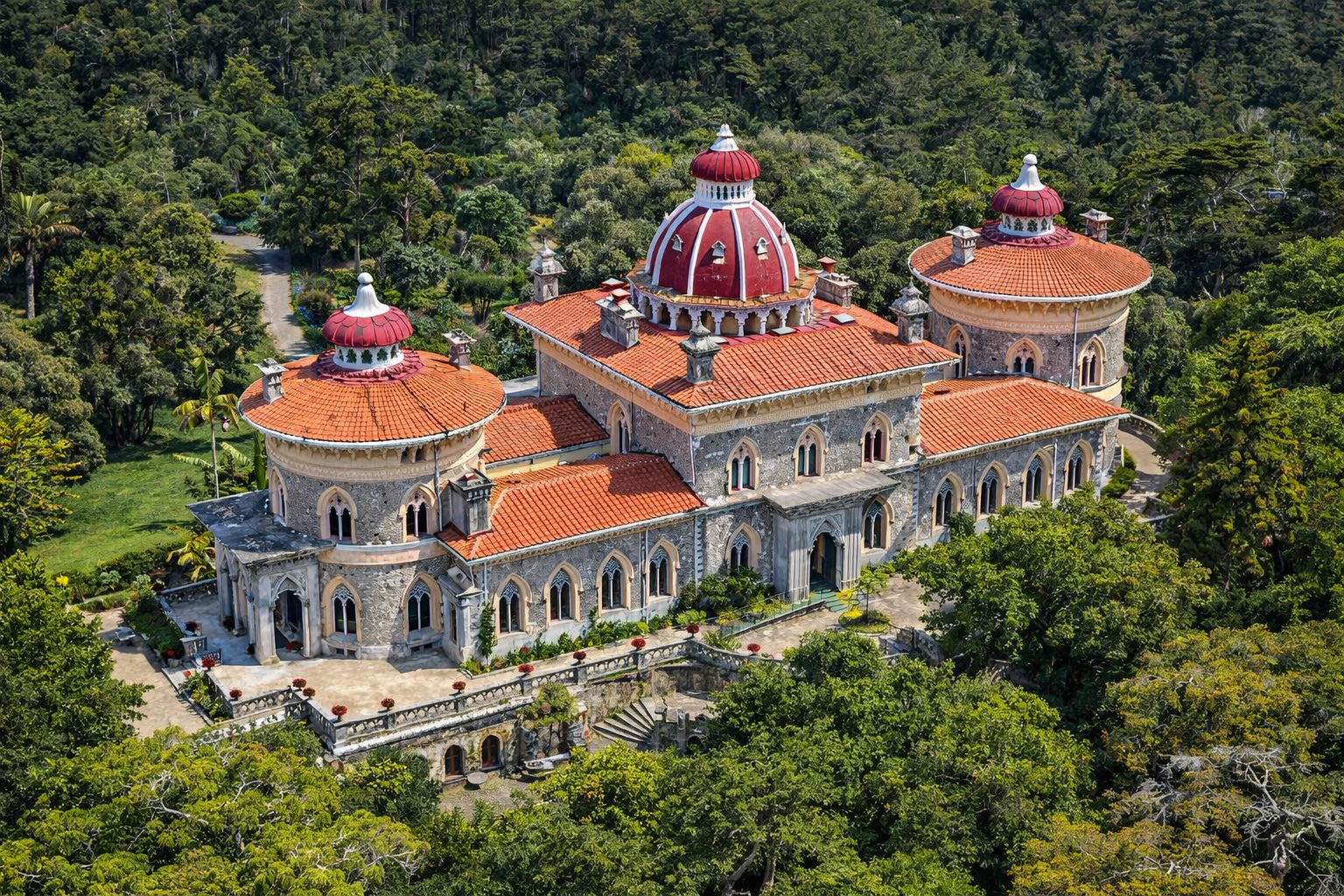 Luftaufnahme des Palácio de Monserrate bei Sintra mit grauer Natursteinfassade, maurisch inspirierten Bögen und drei roten Kuppeln, eingebettet in dichten Parkwald mit Wegen und Gartenflächen