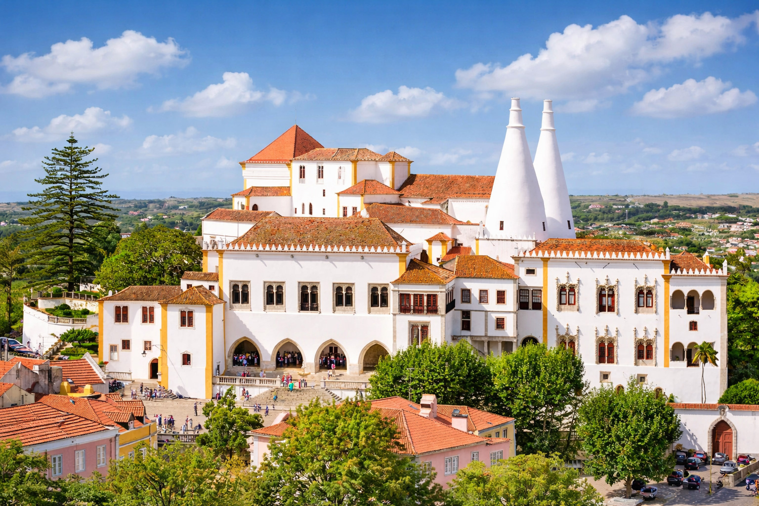 Weißer Königspalast von Sintra mit roten Ziegeldächern und den markanten doppelkegelförmigen Schornsteinen, umgeben von grünen Gärten und der Altstadt vor hügeliger Landschaft unter hellem Himmel