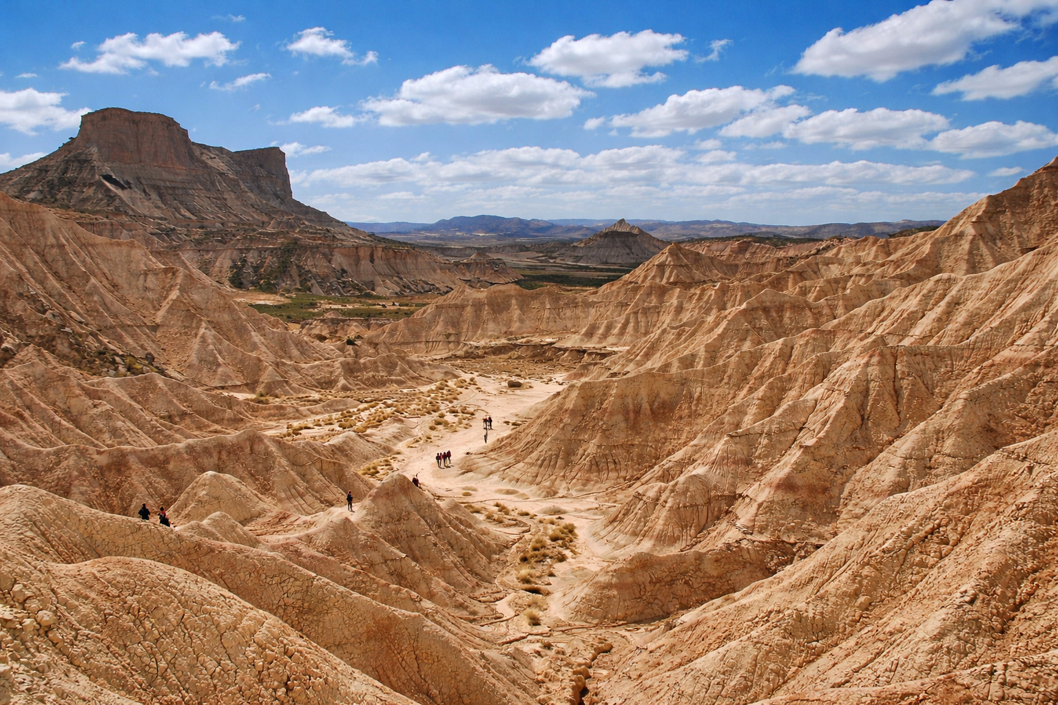 Weitblick über die Bardenas Reales mit hellen Ton- und Sandsteinrücken, tief eingeschnittenen Erosionsrinnen und einer windenden Staubpiste, im Hintergrund Tafelberge und Hügelketten unter leicht veränderten, lockeren Wolken im kräftigen Sonnenlicht