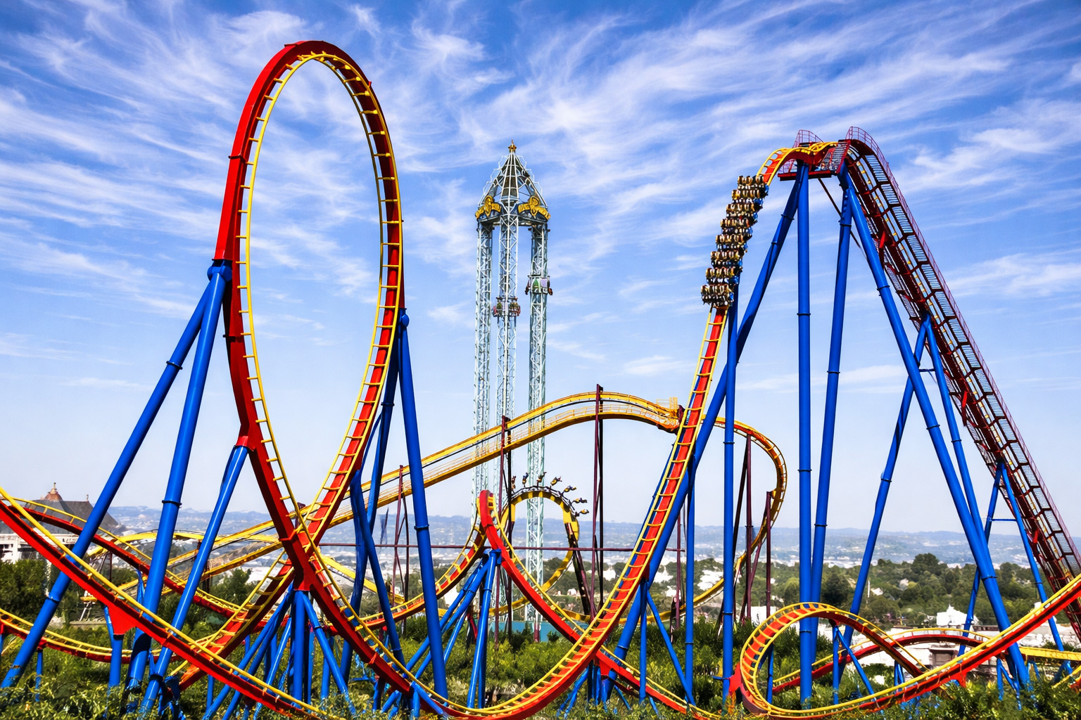 Achterbahnlandschaft im Parque Warner Madrid mit roten und gelben Schienen, hohen Loopings und steilen Abfahrten auf blauen Stahlstützen, im Hintergrund ein Freifallturm und Parkanlagen unter einem Himmel mit feinen, schleierartigen Zirruswolken