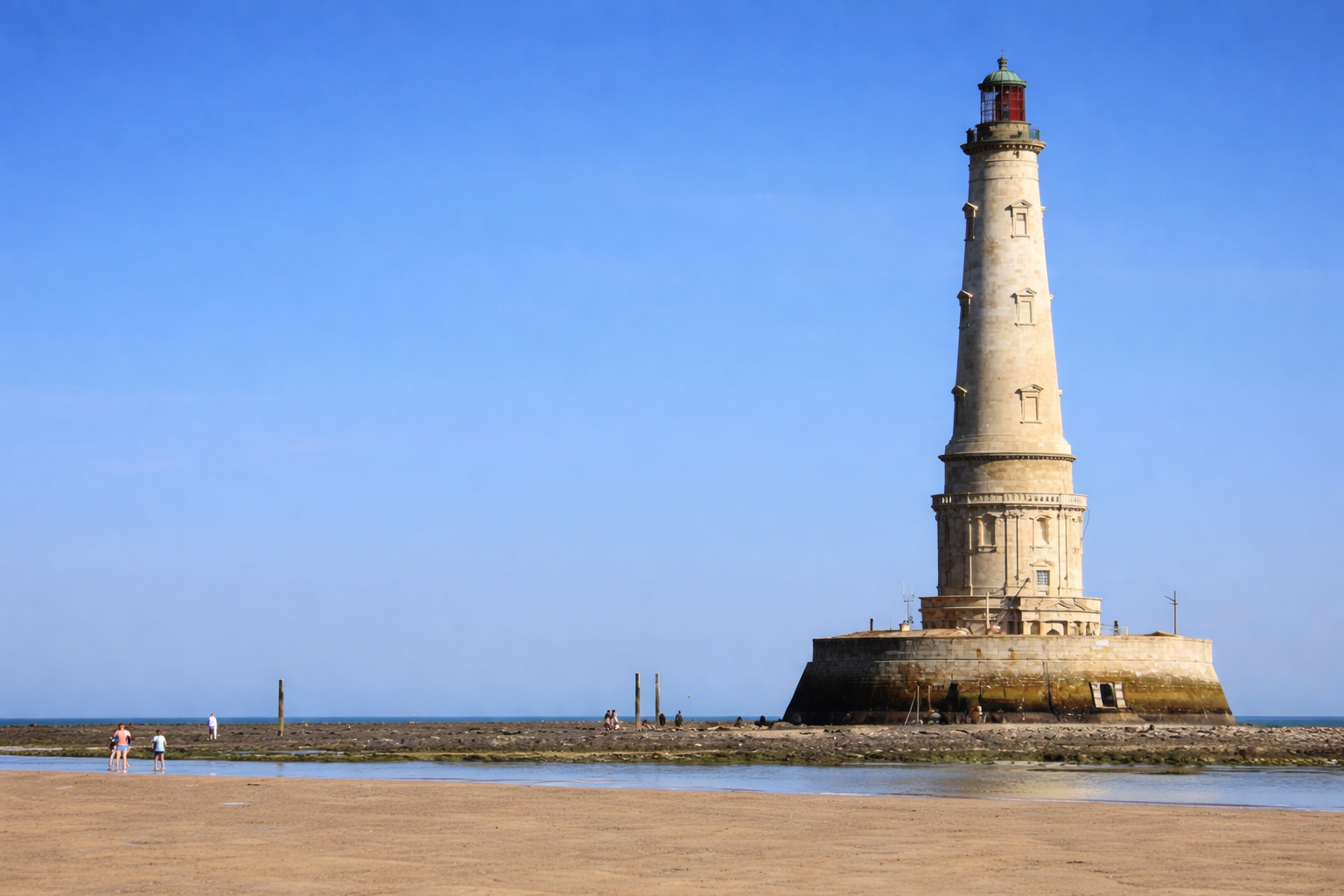 Der Phare de Cordouan ragt als heller Steinturm auf einer Gezeitenbank aus dem Meer, davor breiter Sandstrand und flache Wasserläufe bei Ebbe, im Hintergrund offene See unter klarem blauem Himmel