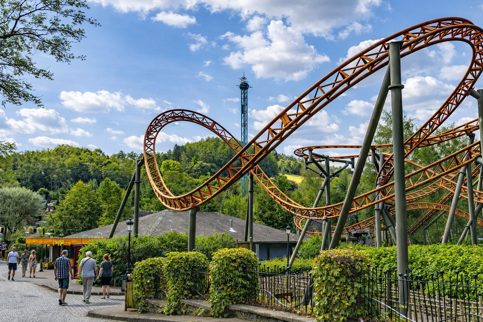 Orangefarbene Achterbahn mit Loopings und geschwungenen Schienen im Plopsaland Ardennes, davor ein gepflasterter Weg mit einigen Spaziergängern links, umgeben von grünen Hecken und Zaunanlagen, im Hintergrund ein hoher Fallturm und bewaldete Hügel unter einem Himmel mit leicht veränderten, lockeren Wolken