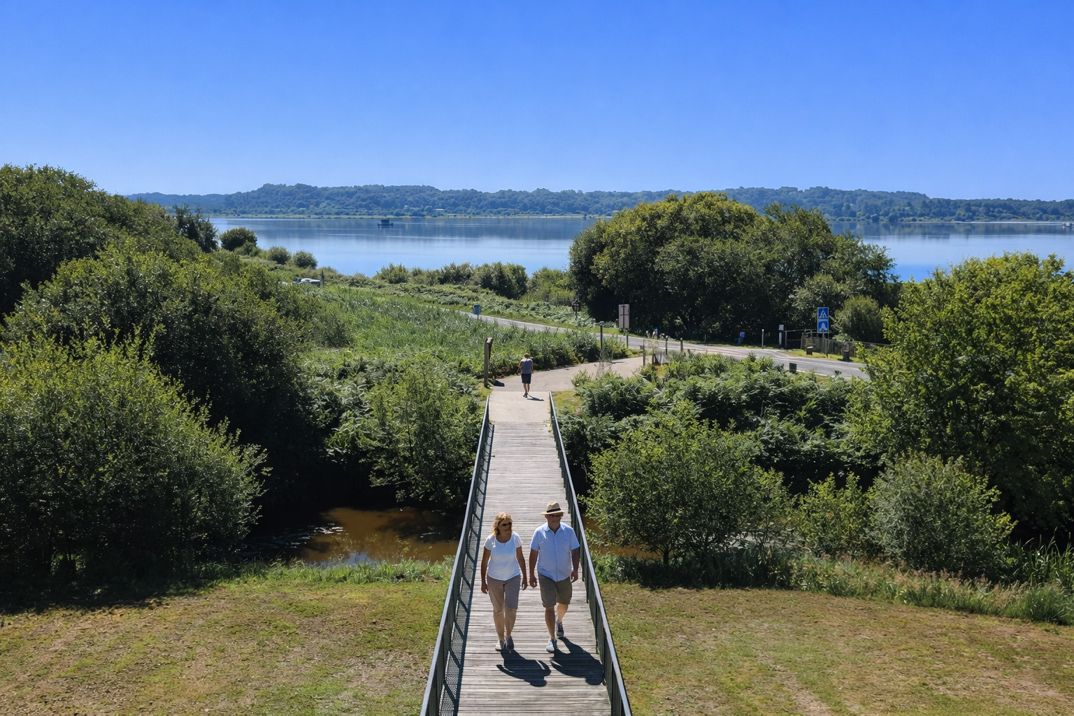 Holzsteg durch die Feuchtlandschaft der Réserve Naturelle du Marais d'Orx, umgeben von dichtem Grün, mit Blick auf einen ruhigen See und bewaldete Ufer; Besucher spazieren über den Steg bei klarem blauem Himmel und kräftigen Schatten