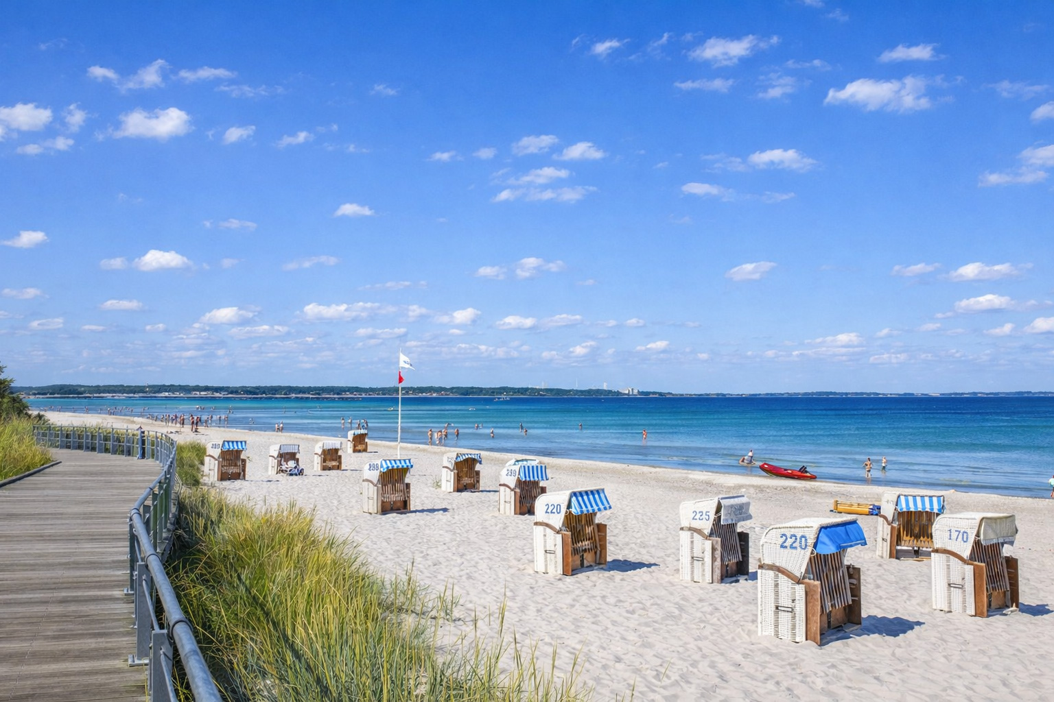 Strand von Scharbeutz mit Dünenpromenade und Blick über helle Sandfläche auf die Ostsee, einzelne Strandkörbe mit blau-weißen Markisen im Vordergrund, ruhiges Wasser und weiter Horizont unter blauem Sommerhimmel