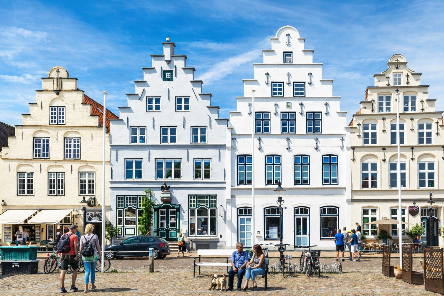 Historische Giebelhäuser am Marktplatz in Schleswig mit weißen und cremefarbenen Fassaden und treppenförmigen Giebeln, davor Kopfsteinpflaster, Fahrräder und eine Sitzbank mit Besuchern, im Vordergrund kleine Straßencafés und Laternen, darüber blauer Himmel mit leicht veränderten Wolken bei kräftigem Sonnenlicht
