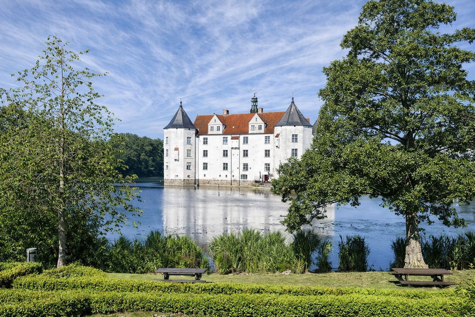 Blick vom Parkufer über den Schlossteich auf Schloss Glücksburg mit weißer Fassade, roten Ziegeldächern und zwei dunklen Ecktürmen, das Schloss spiegelt sich im ruhigen Wasser, im Vordergrund Rasen mit Hecken, Bänken und Schilf, gerahmt von großen Bäumen unter einem Himmel mit feinen, nebligen Zirruswolken bei kräftigem Sonnenlicht