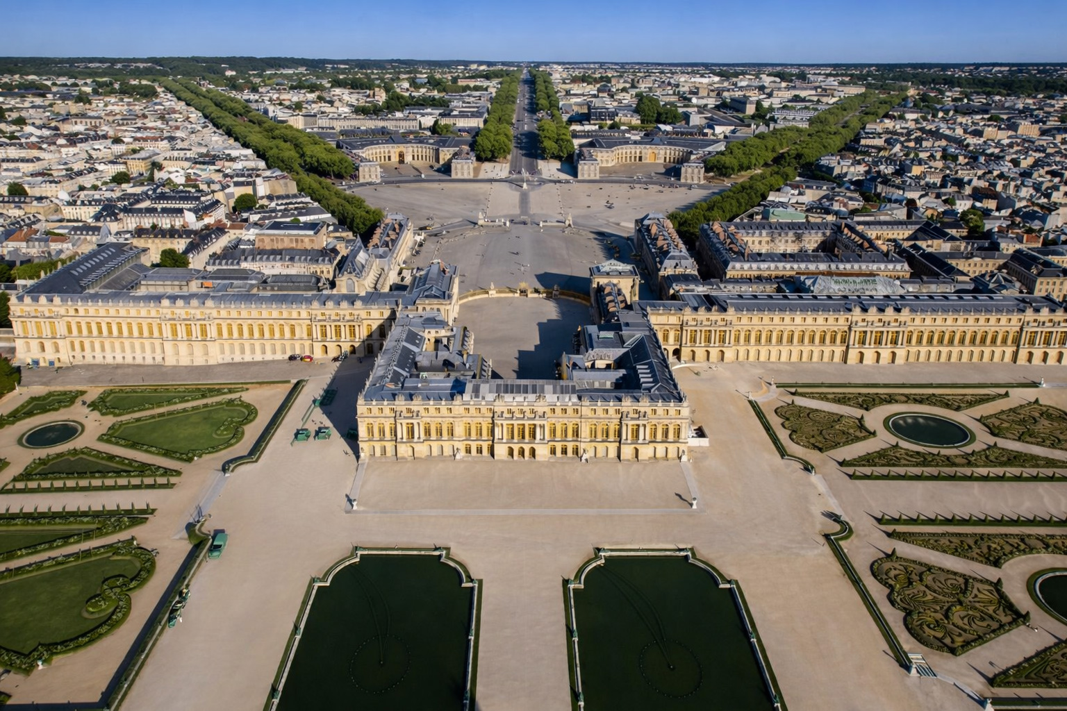 Luftaufnahme des Schlosses Versailles mit weitläufigem Ehrenhof, symmetrischen Flügelbauten und streng angelegten Gartenparterres mit Wasserbecken, dahinter die breite Allee und die Stadt Versailles