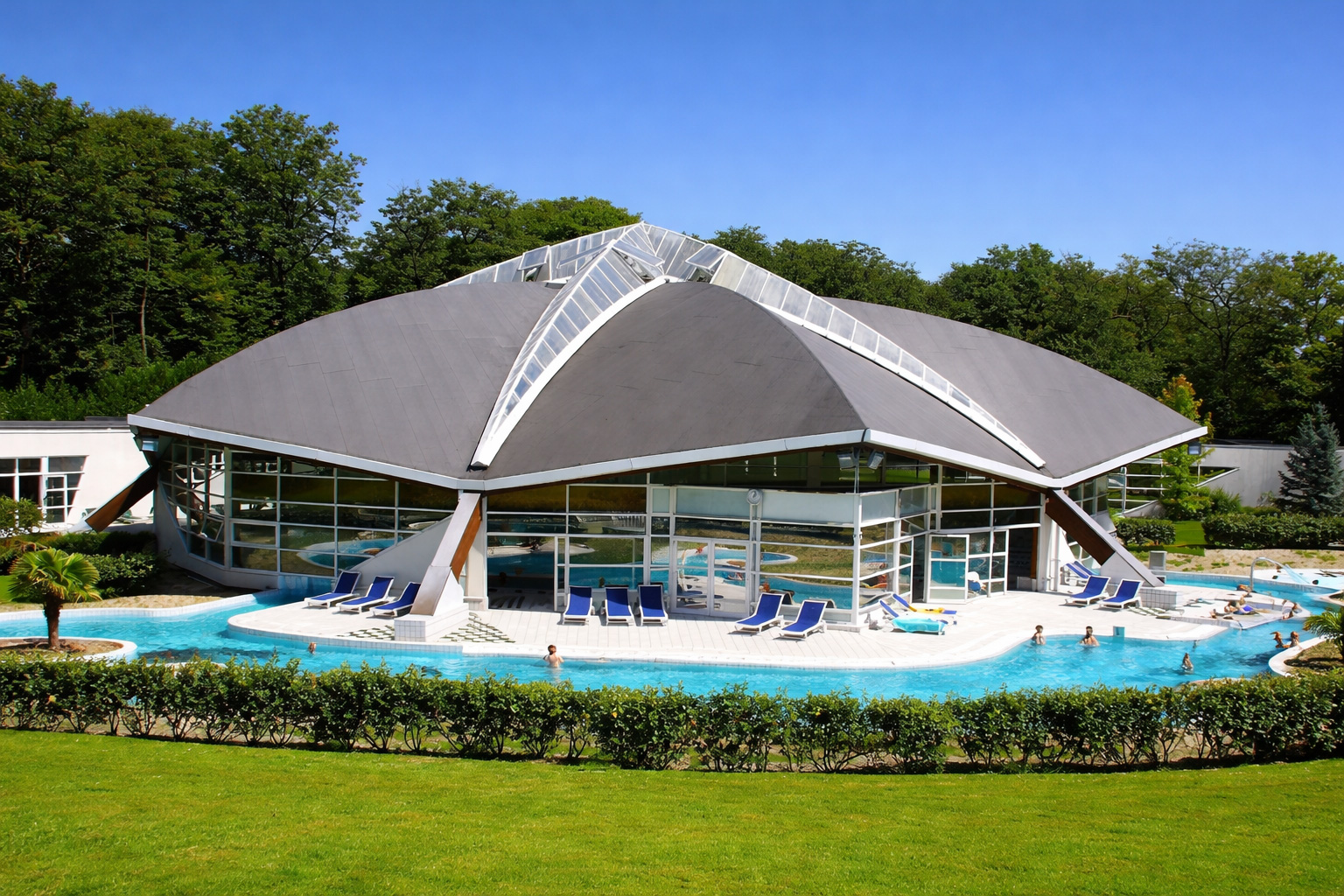 Moderne Thermenanlage in Amnéville mit markantem geschwungenem Dach und großer Glasfront, davor ein türkisblaues Außenbecken mit Liegen, umgeben von gepflegten Hecken und Waldkulisse unter klarem blauem Himmel