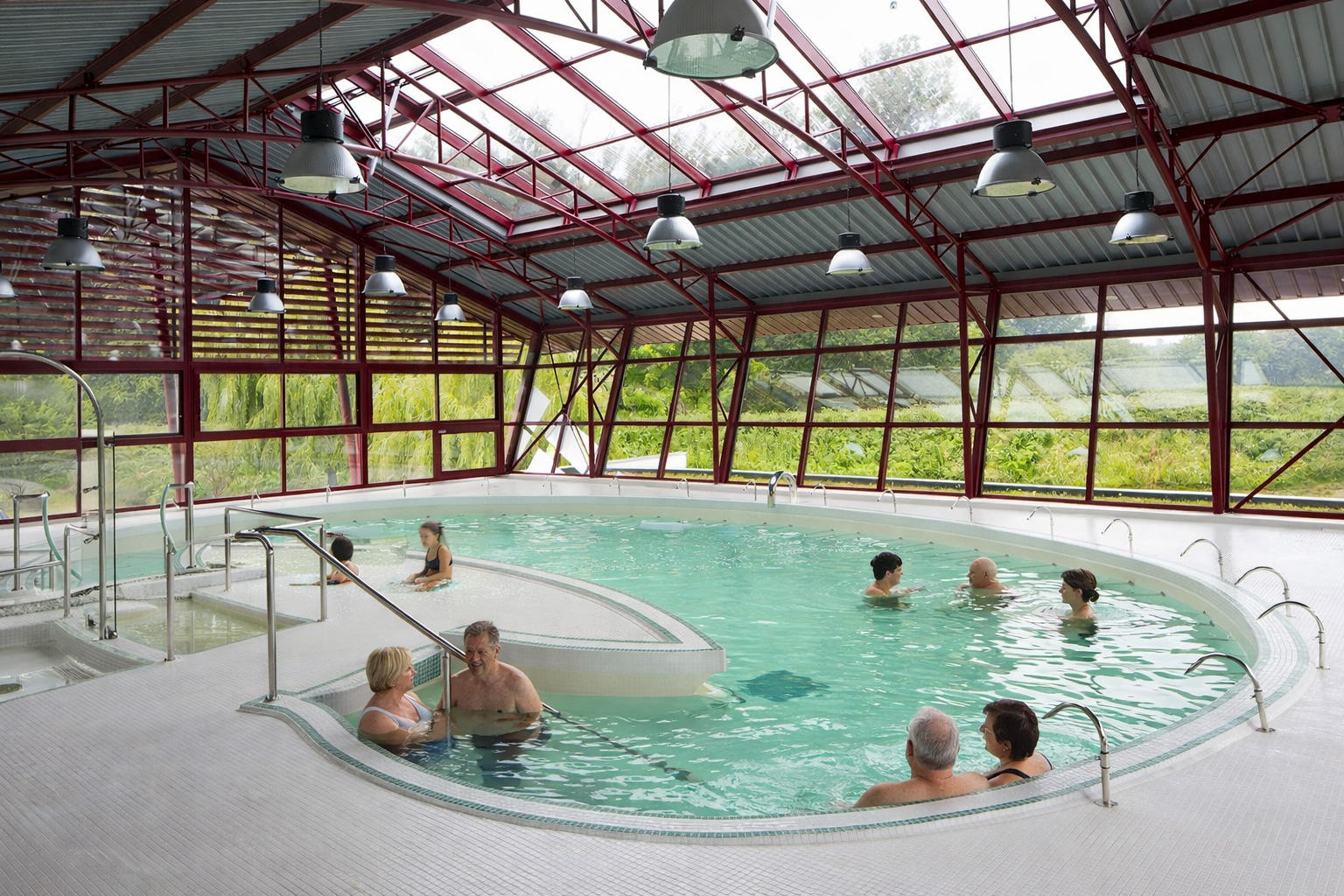 Helles Thermalbad mit großem, ovalem Becken in einer Glas- und Stahlhalle, roten Trägern und weitem Blick ins Grün; mehrere Badegäste entspannen im türkisfarbenen Wasser an Sitzstufen und Geländern