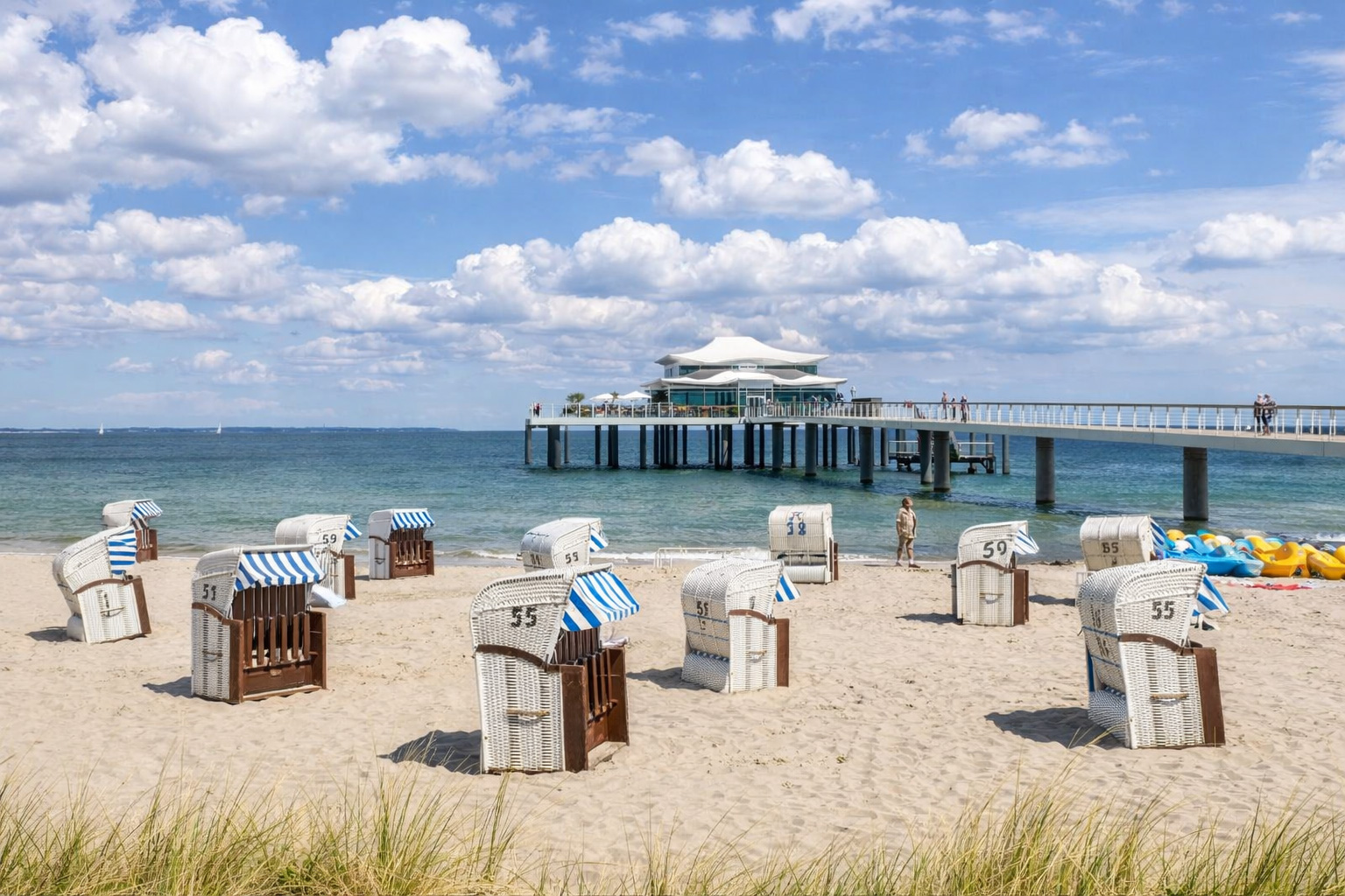 Strand von Timmendorfer Strand mit hellen Strandkörben und blau-weiß gestreiften Markisen auf feinem Sand, lange Seebrücke mit modernem Pavillon über dem Meer, ruhige Ostsee und weiter Himmel über der Küste