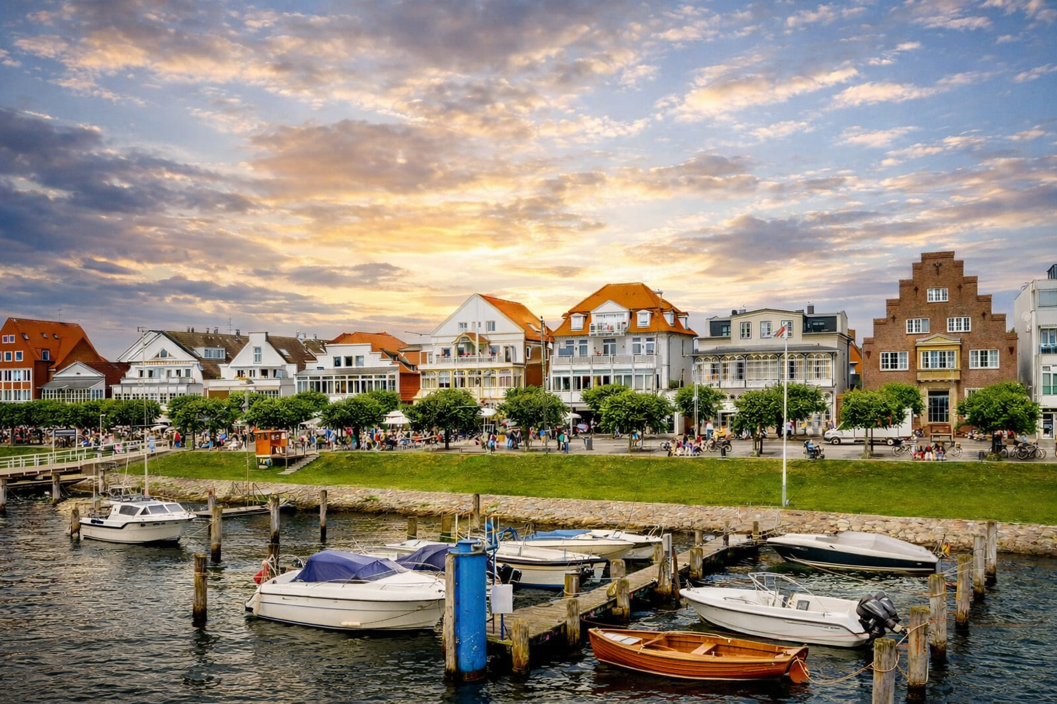 Hafenpromenade in Travemünde mit Segelbooten an Holzstegen, ruhigem Wasser und einer Reihe farbenfroher Gebäude mit roten Dächern, belebte Uferzone vor dramatischem Abendhimmel über der Küstenstadt