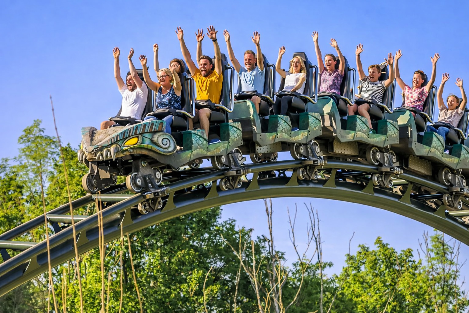 Achterbahnzug im Walibi Belgium auf einer geschwungenen Schiene, vorne ein drachenförmig gestalteter Wagen, Fahrgäste mit erhobenen Armen im hellen Sonnenlicht, darunter grünes Laub und Baumkronen vor blauem Himmel