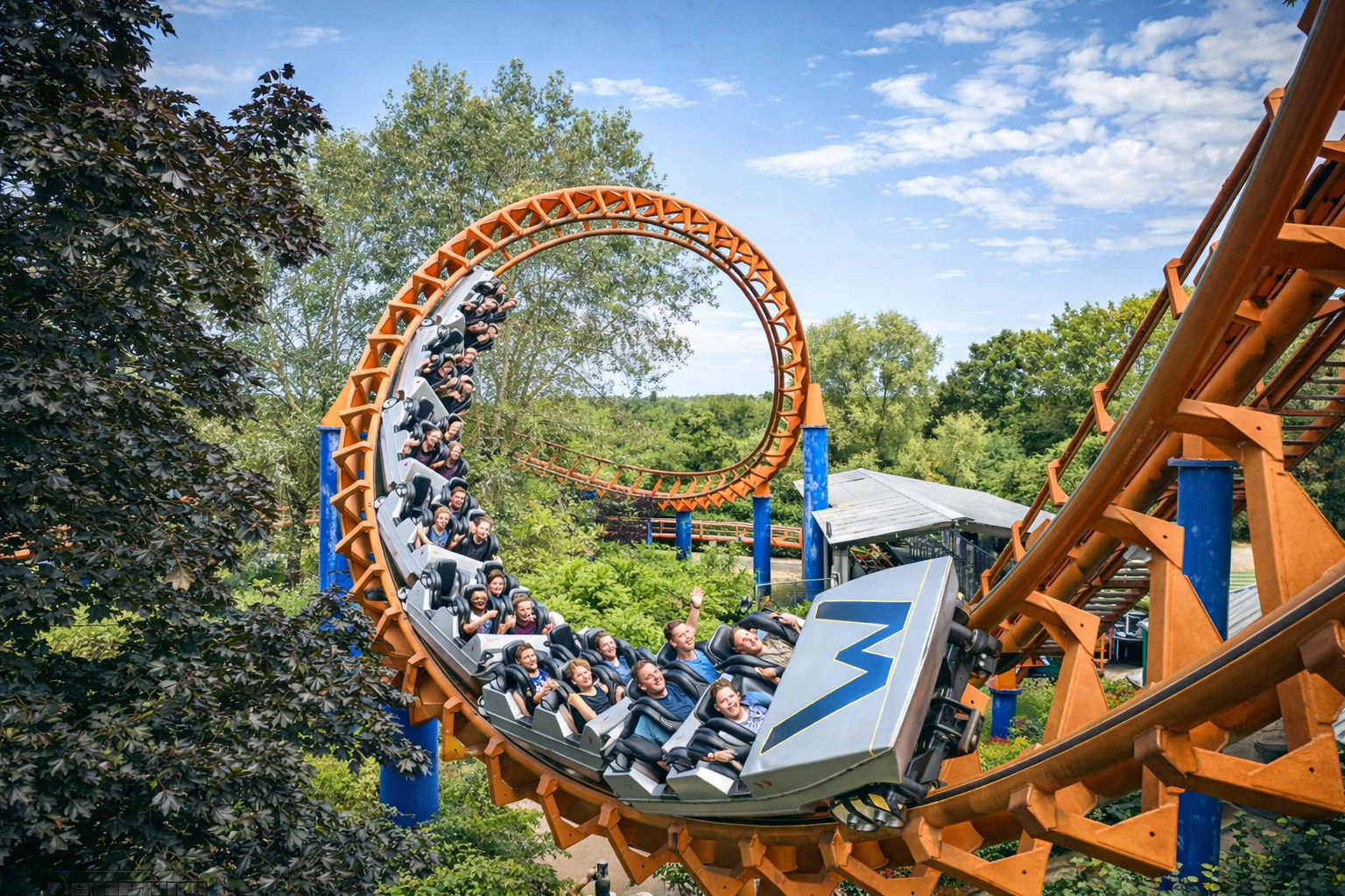 Achterbahnzug in einer engen Loopingkurve auf orangefarbener Stahlstrecke mit blauen Stützen im Freizeitpark Walygator Grand Est, umgeben von Bäumen und Grünflächen unter hellem Himmel mit leicht veränderten Wolken