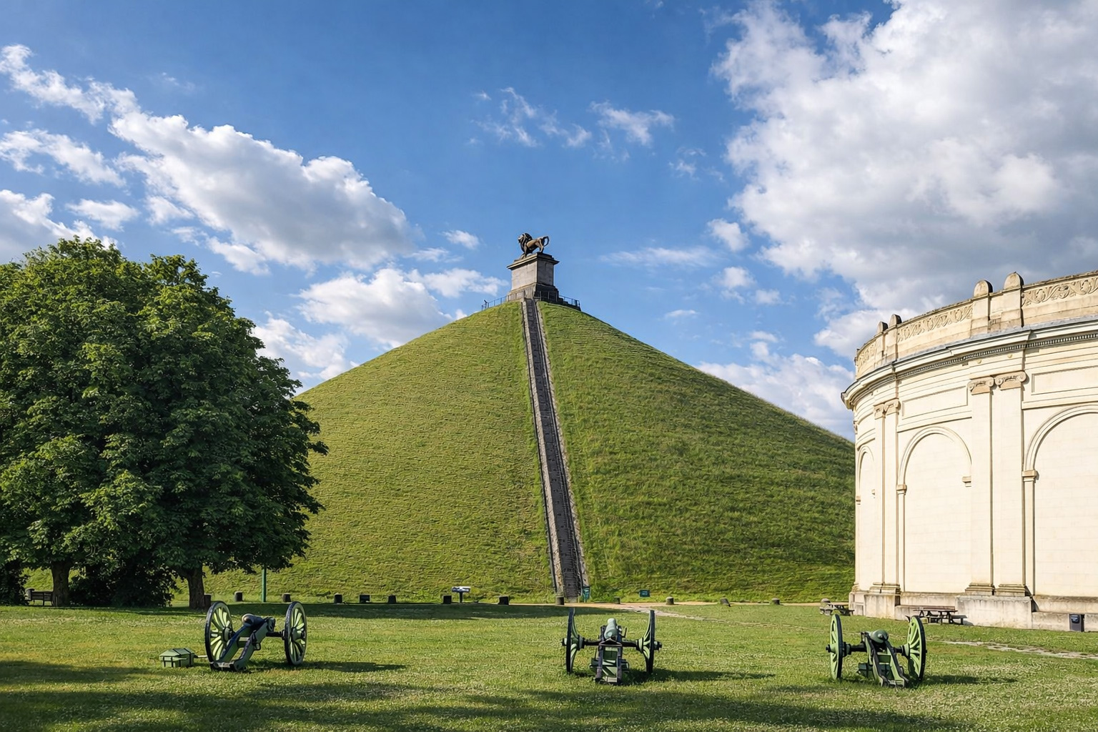 Blick auf den Löwenhügel von Waterloo mit steiler Treppe zum Denkmal und bronzener Löwenstatue auf der Spitze, historische Kanonen im Vordergrund auf grüner Wiese und klassizistisches Museumsgebäude unter blauem Himmel mit markanten Wolken