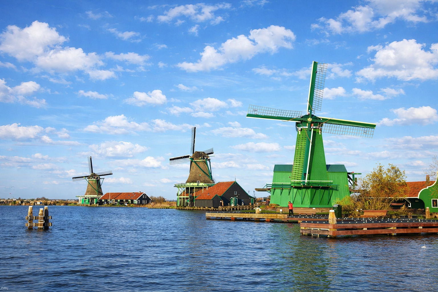 Panorama von Zaanse Schans mit mehreren historischen Windmühlen und grünen Holzbauten am Ufer, ruhiges Wasser im Vordergrund und typisch niederländische Dächer im Hintergrund, aufgenommen bei klarem Sonnenlicht unter einem blauen Himmel mit leicht veränderten, lockeren Wolken
