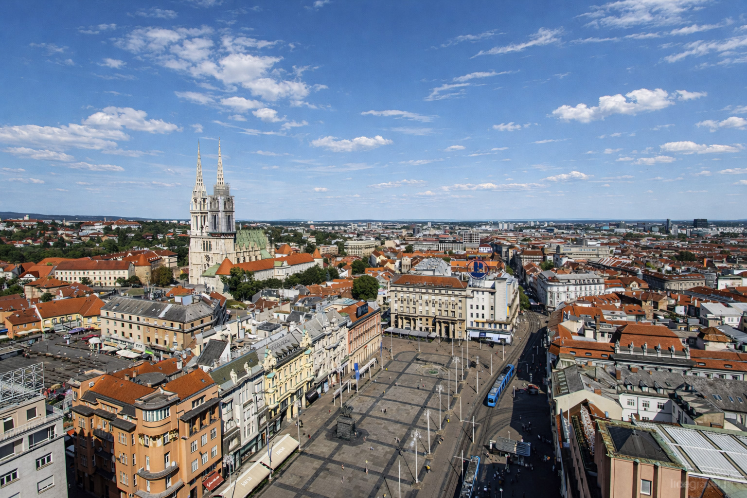 Panoramablick vom Zagreb 360° observation deck über den Ban-Jelačić-Platz im Zentrum von Zagreb mit Straßenbahn, historischen Fassaden, roten Dächern und der Kathedrale von Zagreb unter blauem Himmel mit lockeren Wolken.