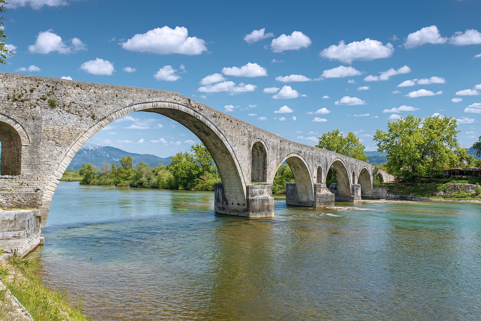 Antike Steinbrücke von Arta in Griechenland mit ihren eleganten Bögen über dem Fluss, umgeben von Bäumen und Bergen im Hintergrund unter blauem Himmel mit weißen Wolken.