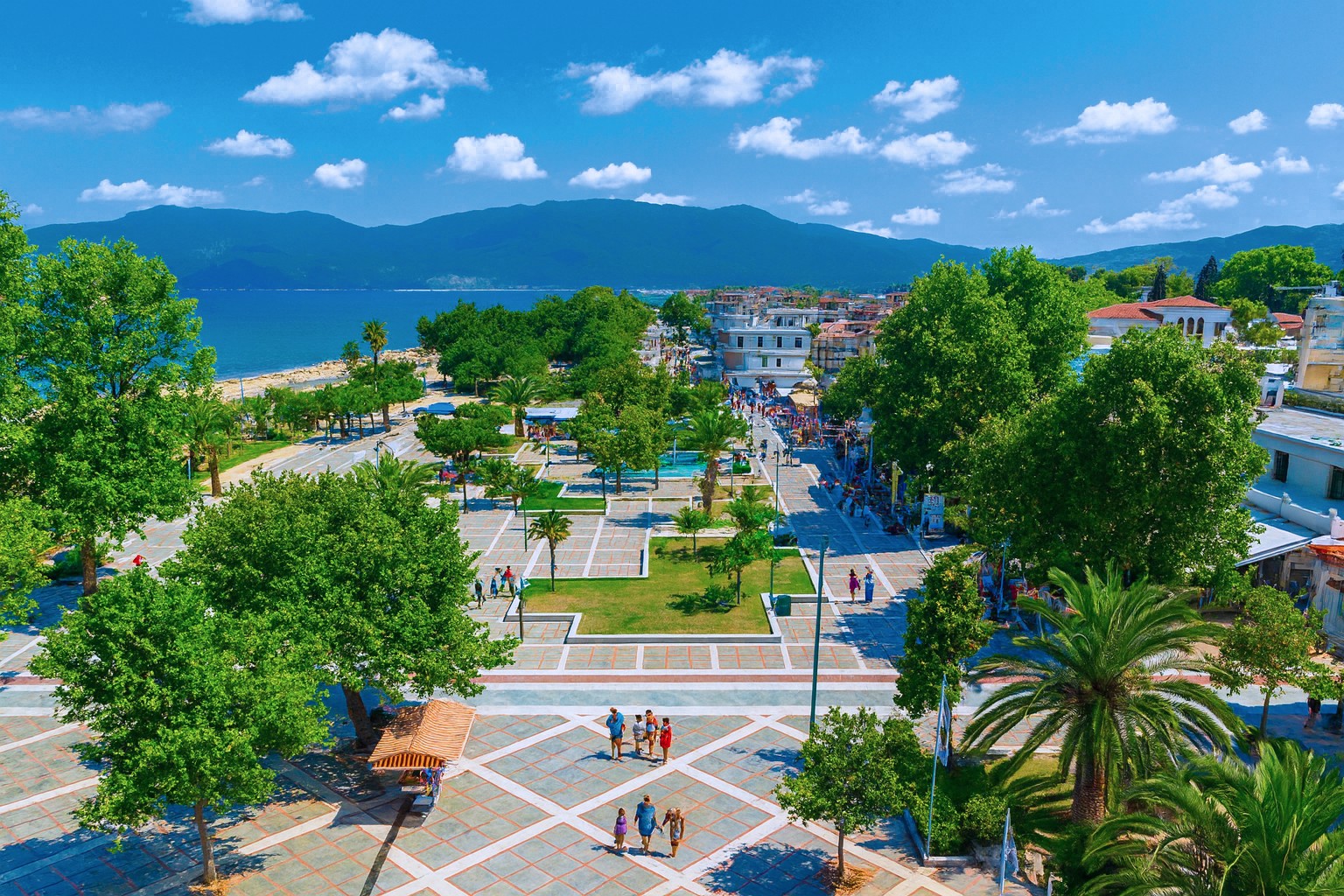 Panorama der Promenade in Asprovalta mit grünen Bäumen, Strand, Meer und Spaziergängern bei sonnigem Himmel mit weißen Wolken.