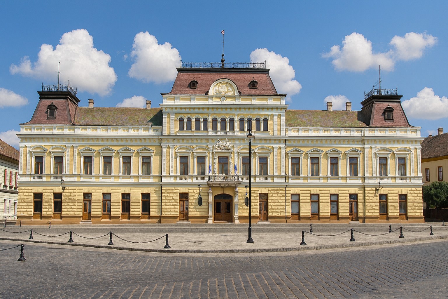Rathaus von Baja in Ungarn mit neoklassizistischer Fassade auf einem freien Platz bei blauem Himmel mit weißen Wolken.