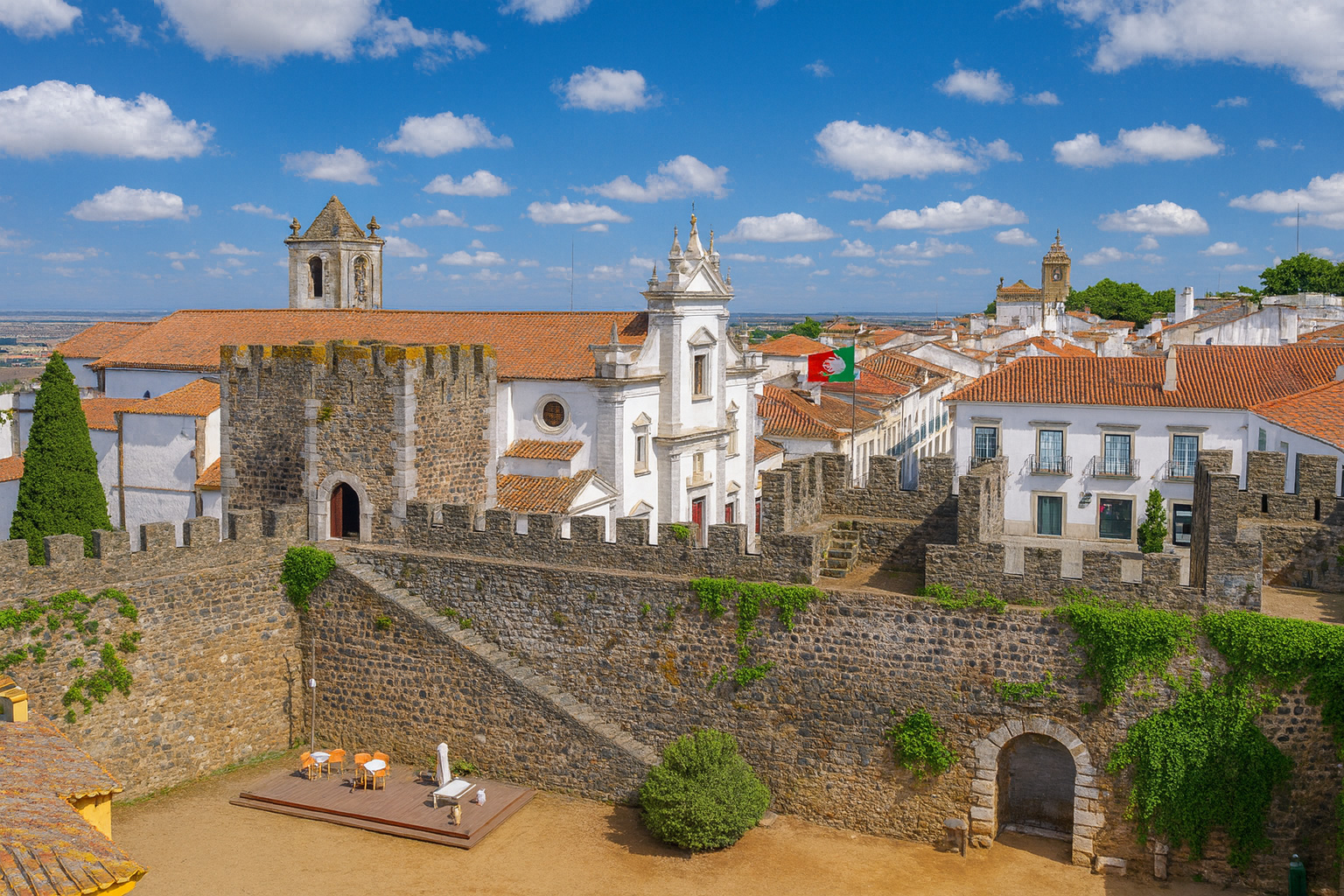 Panoramaaufnahme von der Festung in Beja mit Blick auf die Kirche Sé de Beja und die Altstadt, bei klarem Sonnenlicht und malerischen weiß-grauen Wolken am Himmel.