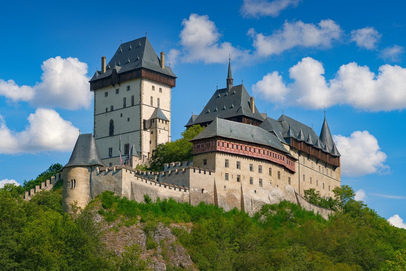 Burg Karlštejn bei Beroun in Tschechien, auf einem Hügel gelegen, bei strahlender Mittagssonne mit malerischen weißen Wolken am blauen Himmel.