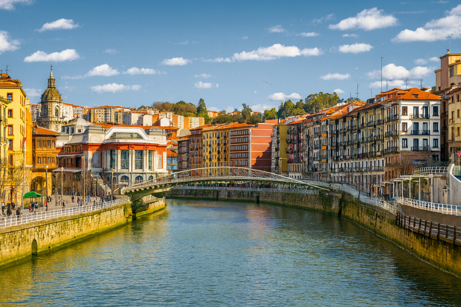 Panorama von Bilbao mit der Zubizuri-Fußgängerbrücke über den Nervión-Fluss, umgeben von historischen Gebäuden und sonnigem Himmel mit weißen Wolken.