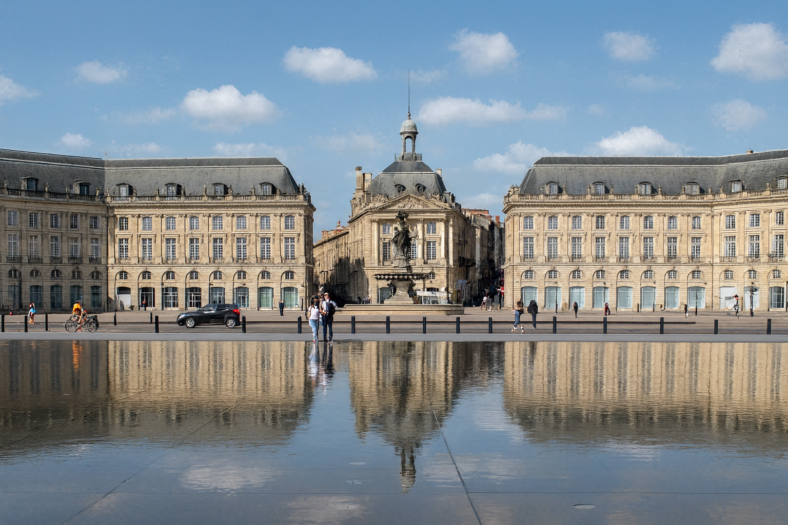 Der Place de la Bourse in Bordeaux mit dem Musée National des Douanes im Hintergrund, klar reflektiert im Miroir d’eau unter blauem Himmel mit weißen Wolken.