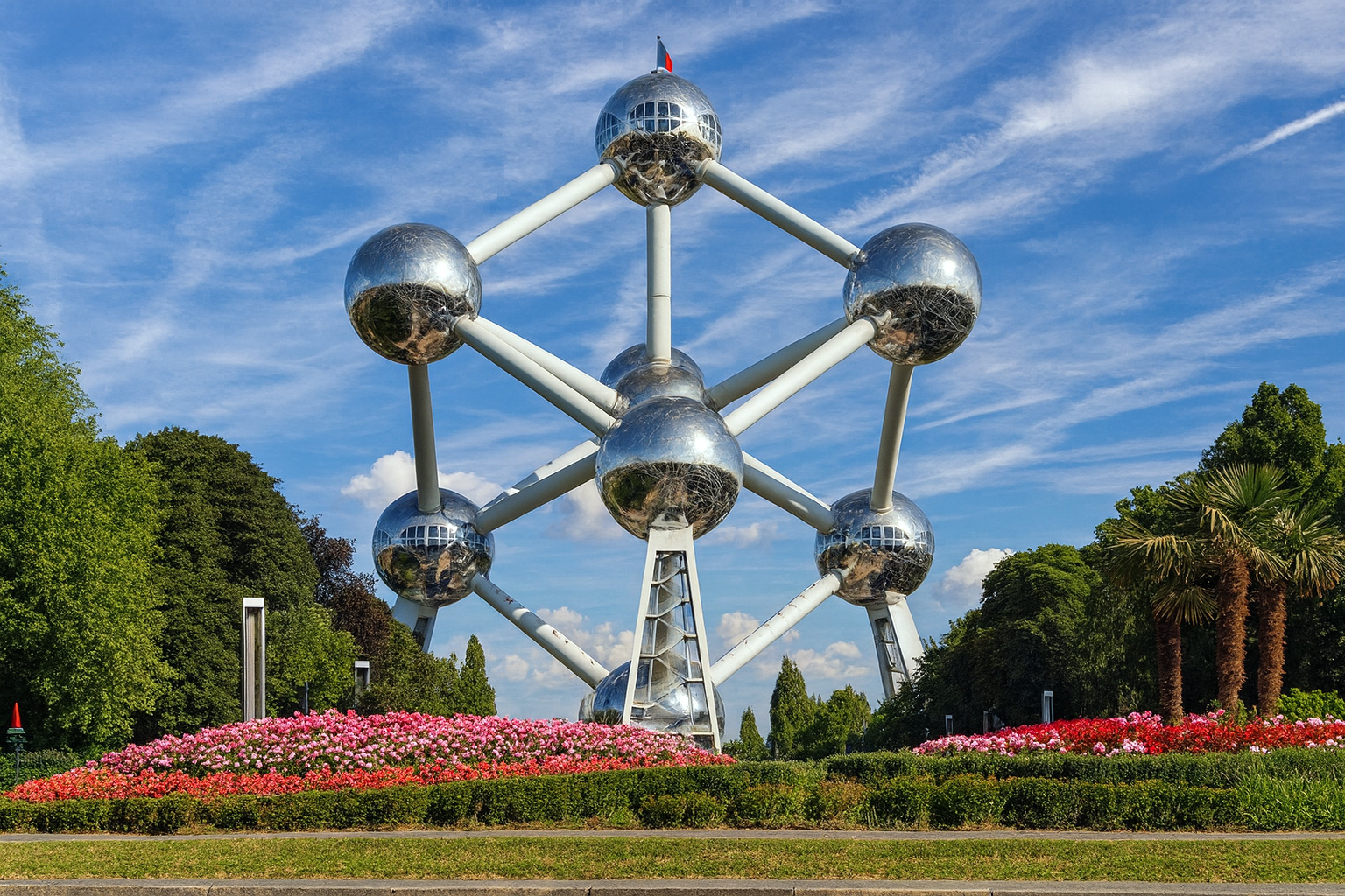 Panorama des Atomium in Brüssel mit reflektierenden Metallkugeln, blühenden Blumenbeeten im Vordergrund und einem Himmel mit Zirrus- und Cumuluswolken bei Sonnenlicht.