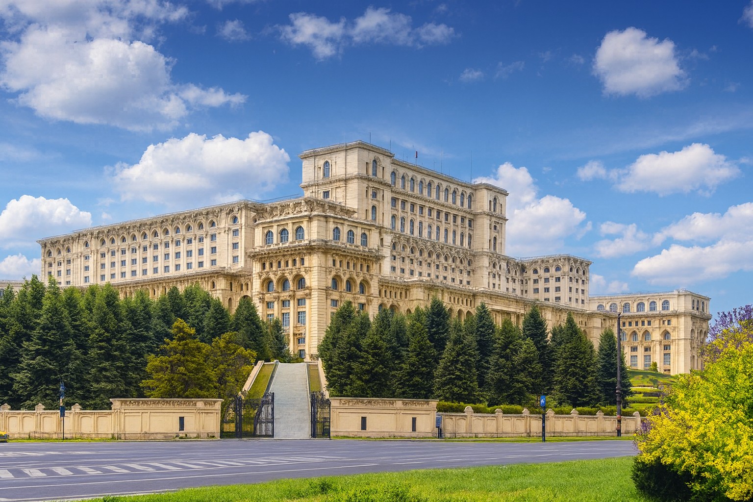 Parlamentsgebäude in Bukarest mit imposanter Fassade, umgeben von Bäumen und blauem Himmel mit weißen Wolken.