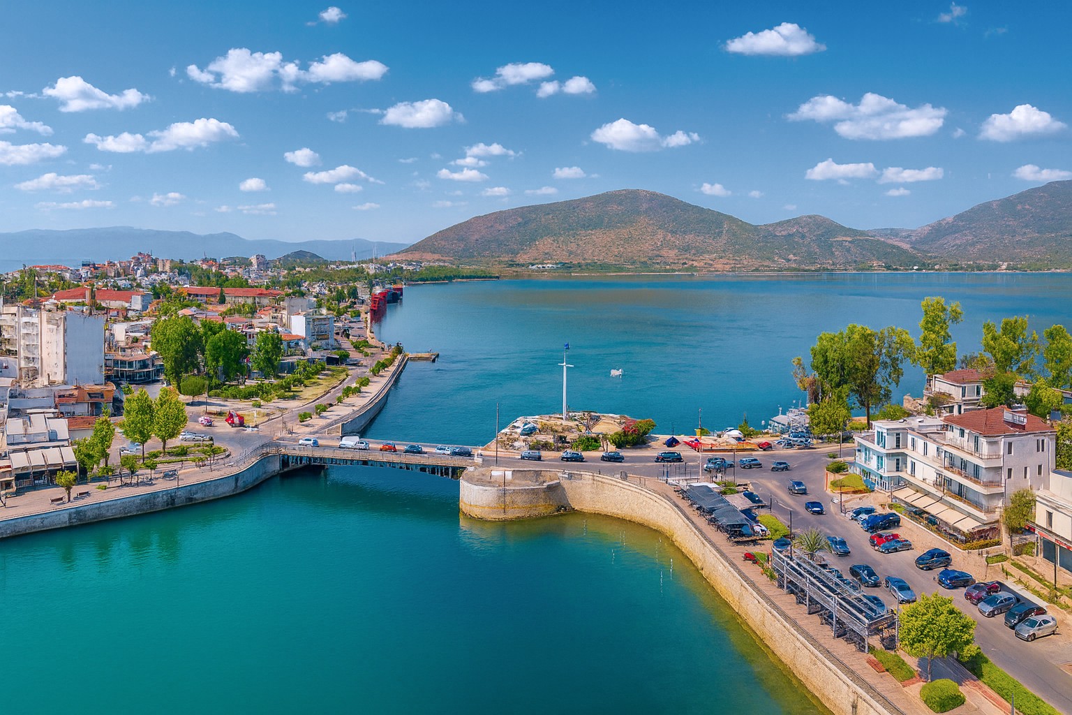 Panorama von Chalkida mit Brücke, Hafenpromenade, parkenden Autos und Blick auf das Meer und die Berge unter blauem Himmel mit weißen Wolken.