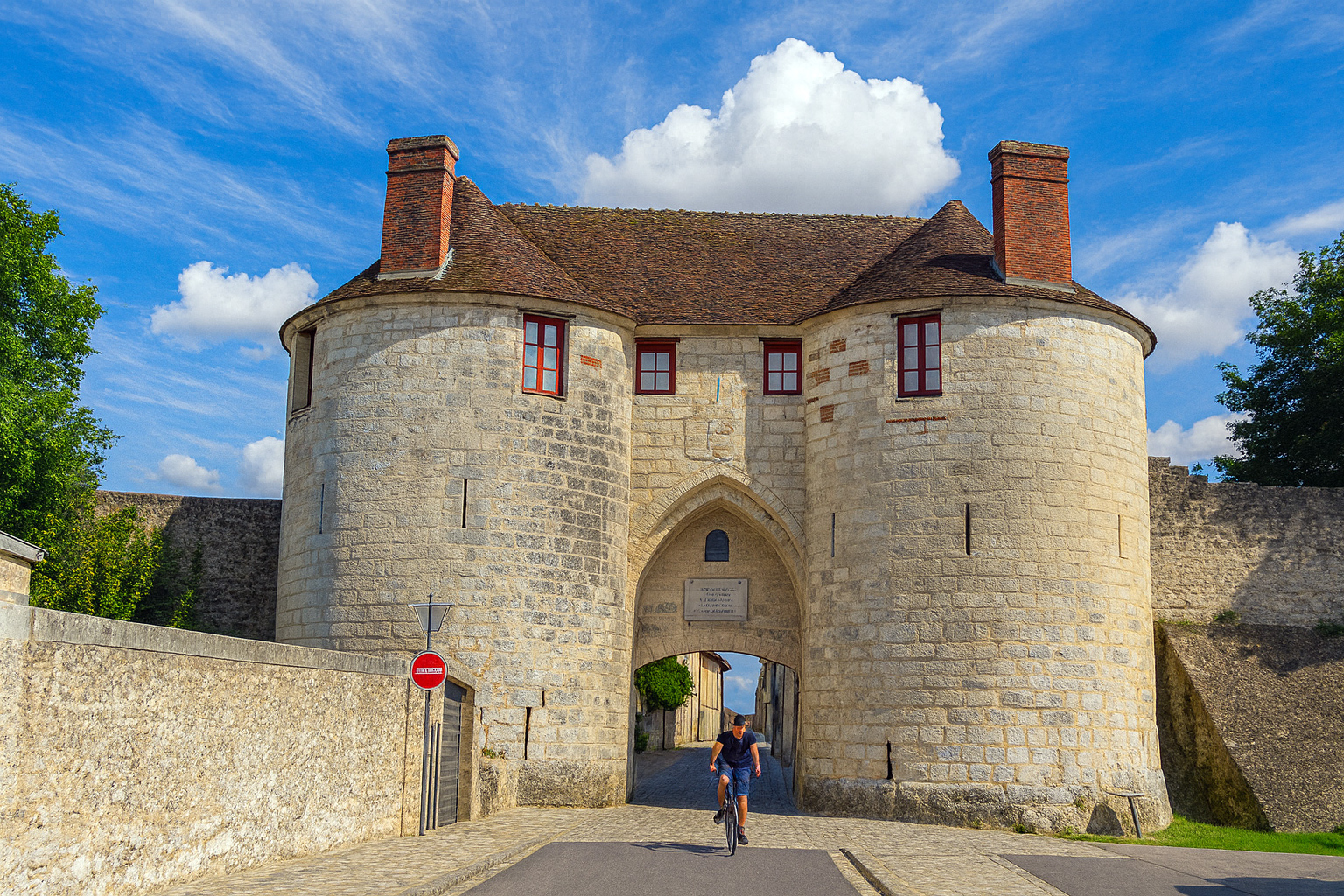 Historisches Stadttor Porte Saint-Pierre in Château-Thierry mit Radfahrer, der aus dem Tor fährt, unter einem Himmel mit Zirrus- und Cumuluswolken bei Sonnenlicht.