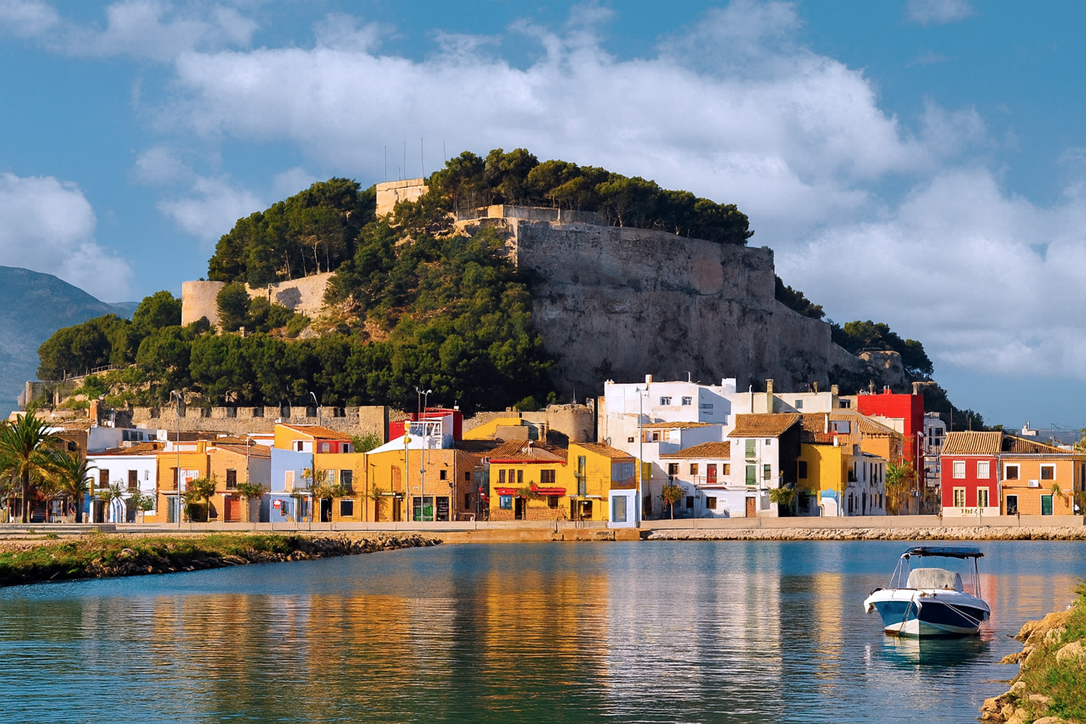 Panoramablick auf die Burg von Dénia mit bunten Häusern und spiegelndem Wasser unter malerischen weiß-grauen Wolken im Sonnenlicht.